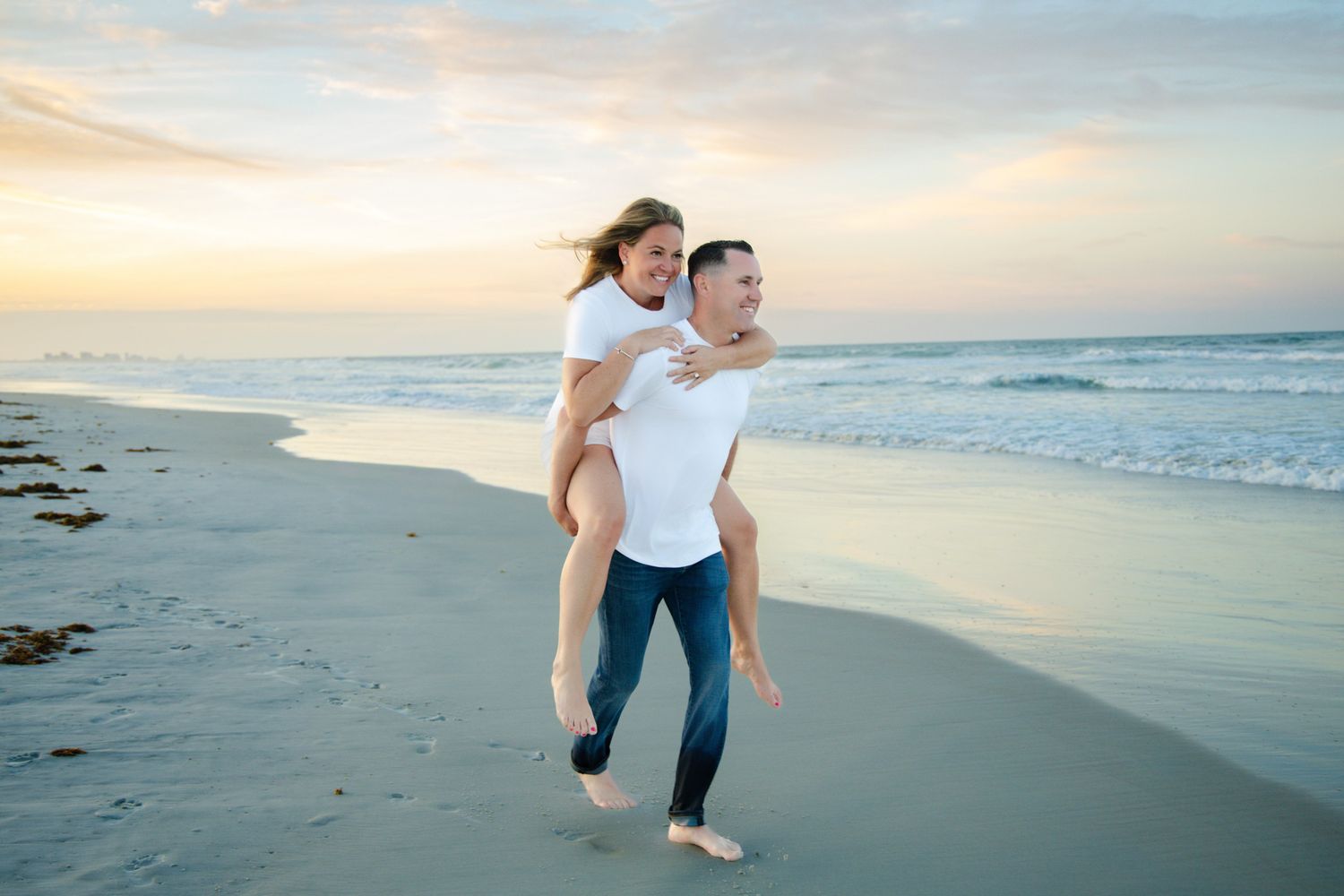 A couple enjoys a romantic piggyback ride on a sandy beach at sunset with ocean waves in the background.