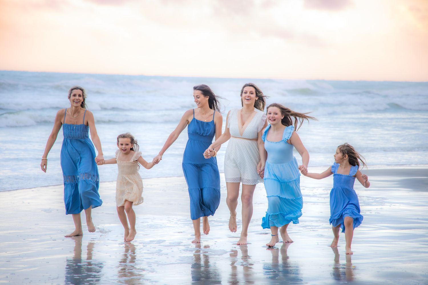 Six women and girls in blue and white dresses run joyfully along a sandy beach at sunset, holding hands.