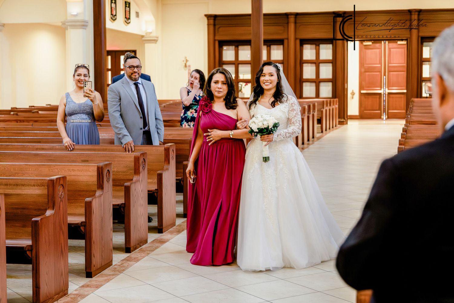 Bride in white gown walks down church aisle with bridesmaid in red dress during wedding ceremony.