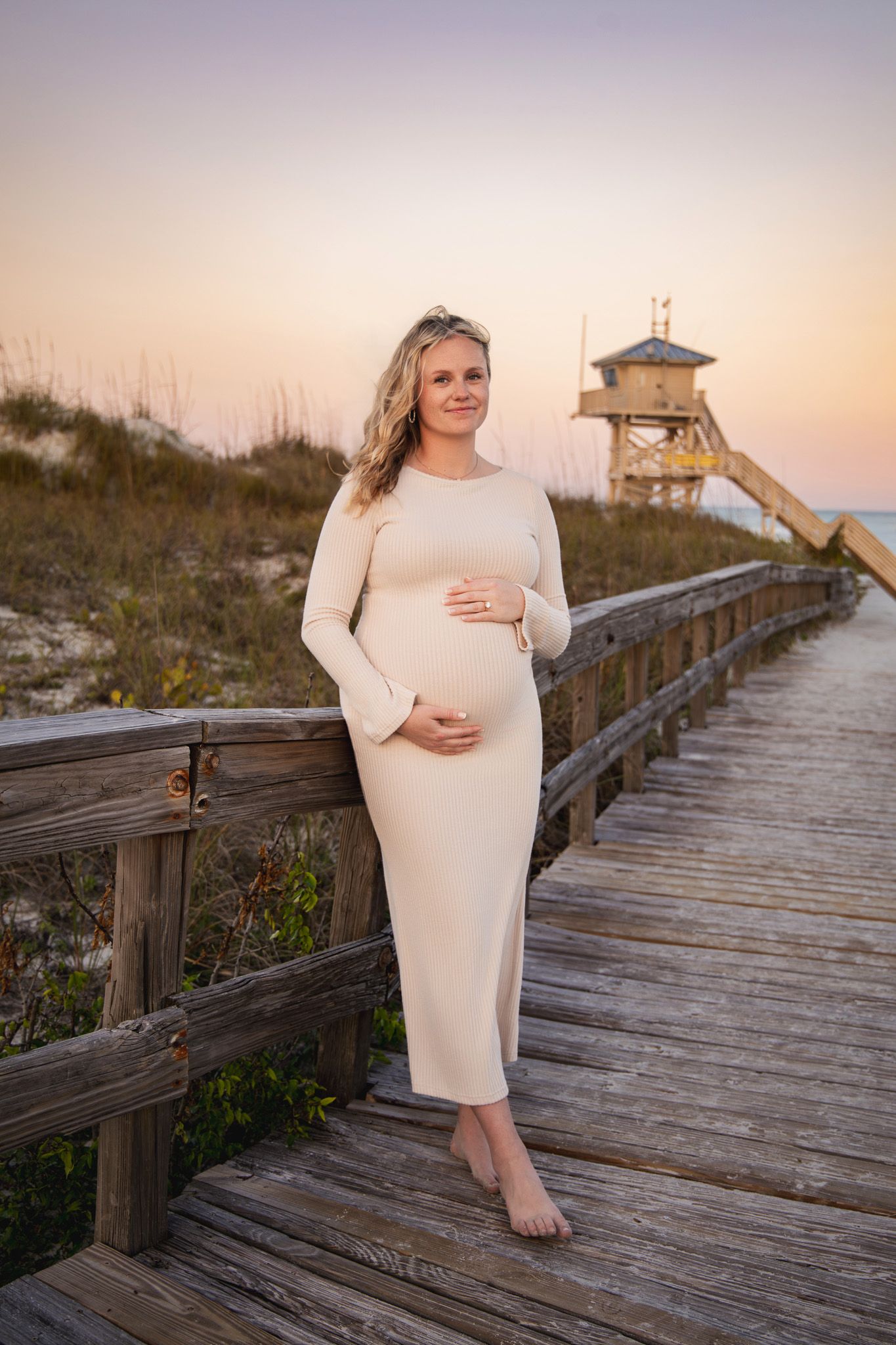A maternity photoshoot on a wooden beach boardwalk at sunset shows a woman in a cream-colored dress cradling her belly.