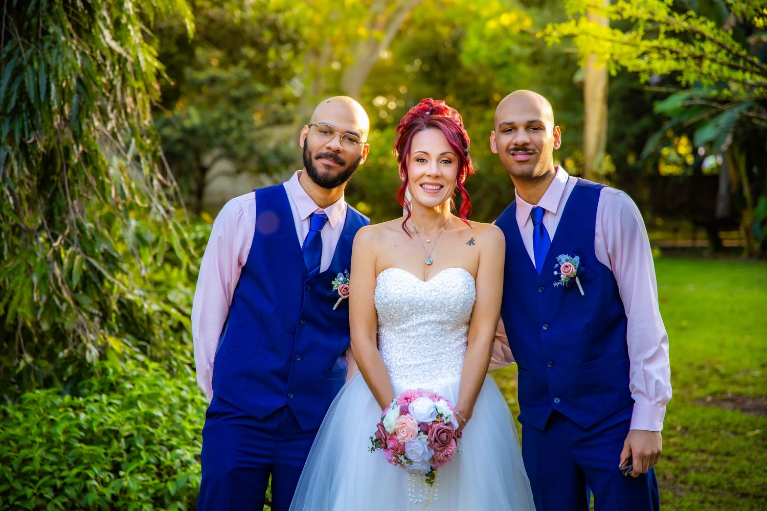 Three people in formal wedding attire pose together in a garden setting with blue vests and a white dress.