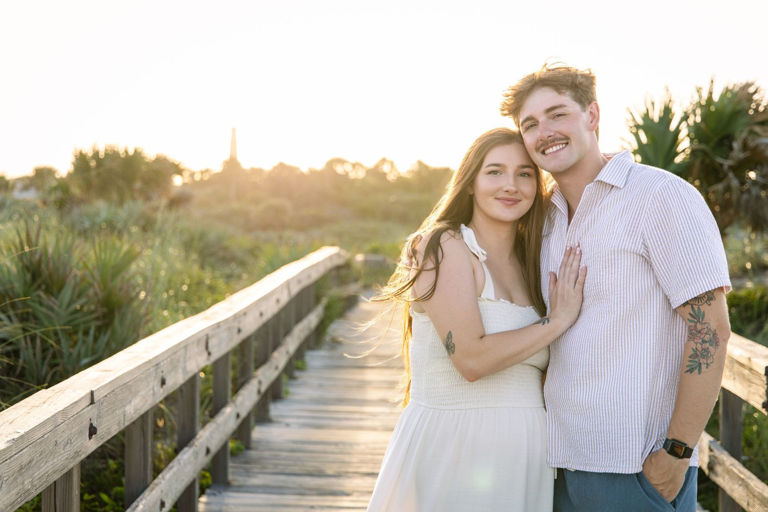Couple posing on a wooden boardwalk at golden hour, surrounded by lush greenery, woman in white dress, man with tattoos.