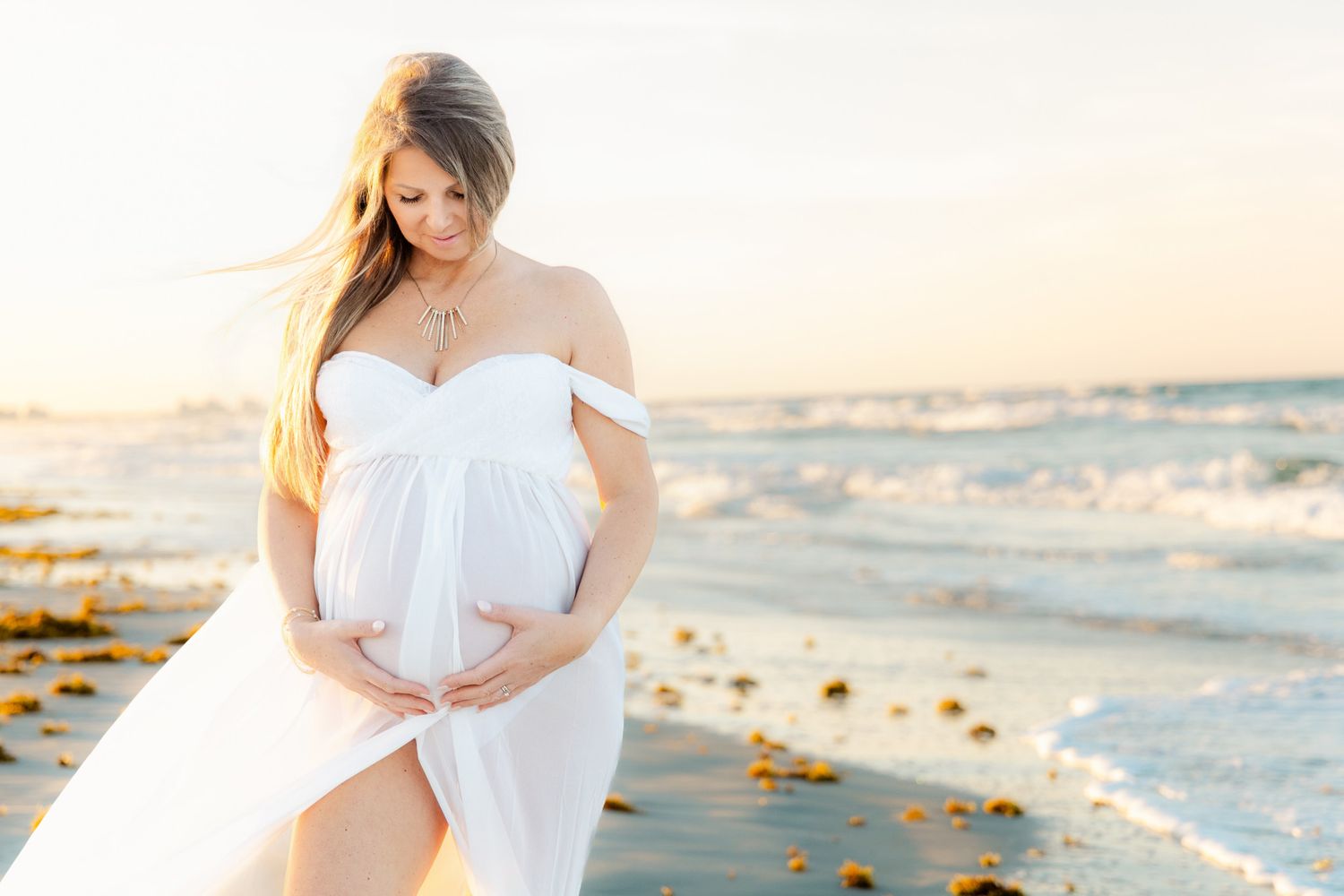 Pregnant woman in white off-shoulder flowing dress cradling baby bump on a sunlit beach with ocean waves.