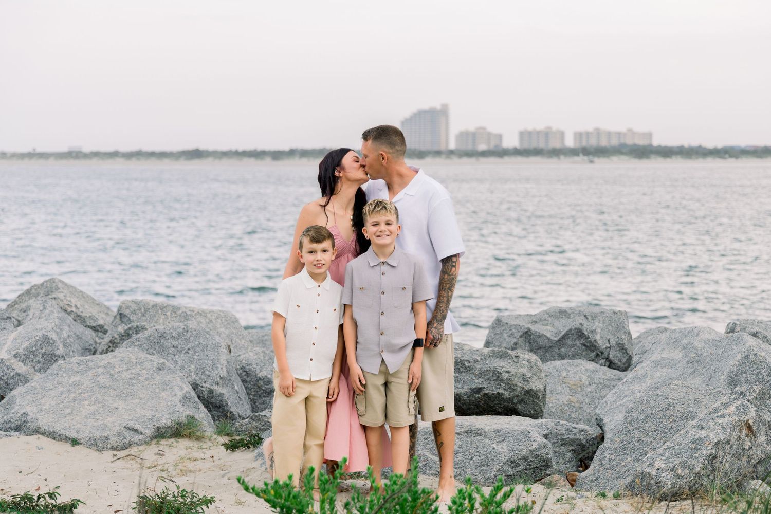 Family of four posing on rocky beach with ocean and city skyline in background on overcast day.