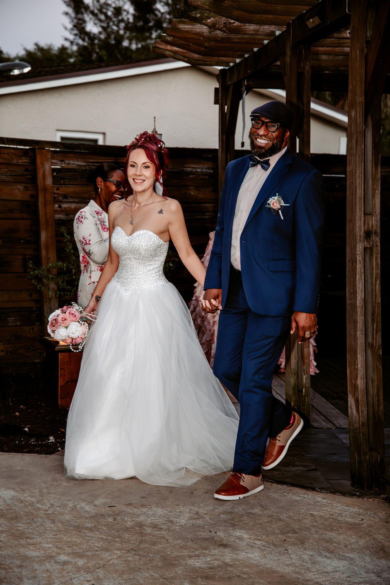A bride in a white ballgown and groom in navy suit show off their matching red sneakers during a rustic wedding photo.