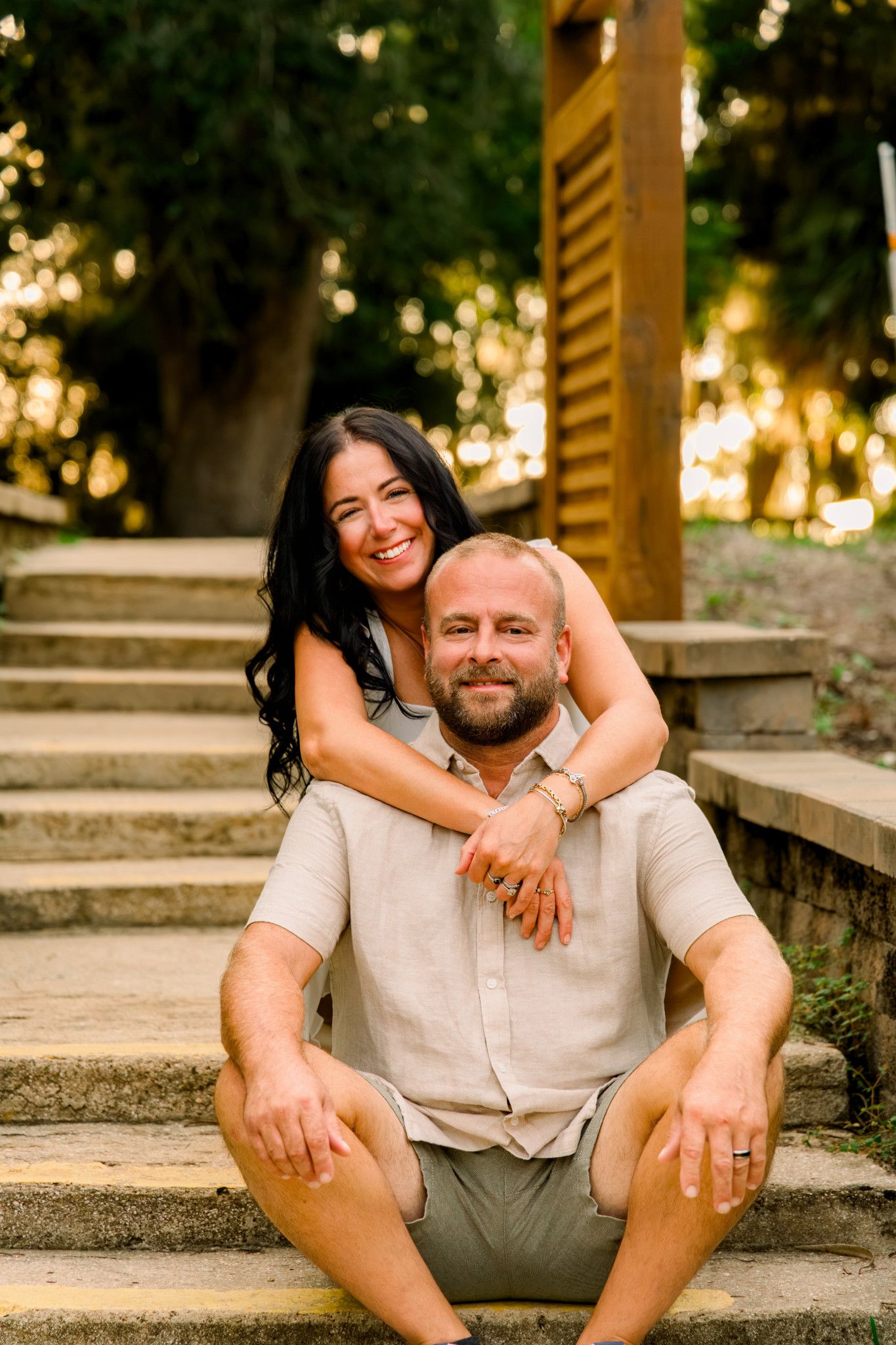 A smiling couple sits on stone steps outdoors, woman embracing man from behind, surrounded by lush green trees.