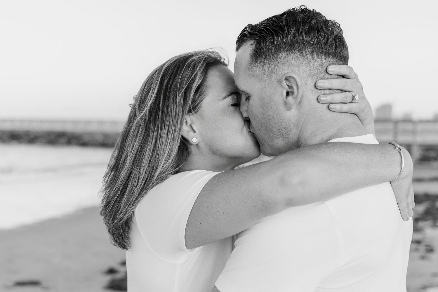 Black and white photo of couple sharing a romantic kiss on the beach, woman embracing mans neck.