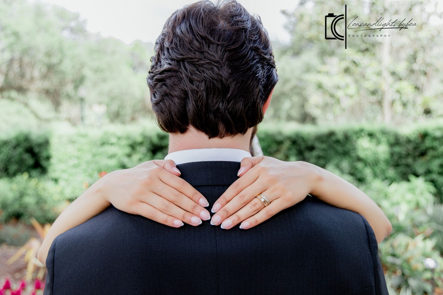 Bride resting hands on groom's shoulders from behind, showing engagement ring, with lush green garden backdrop.