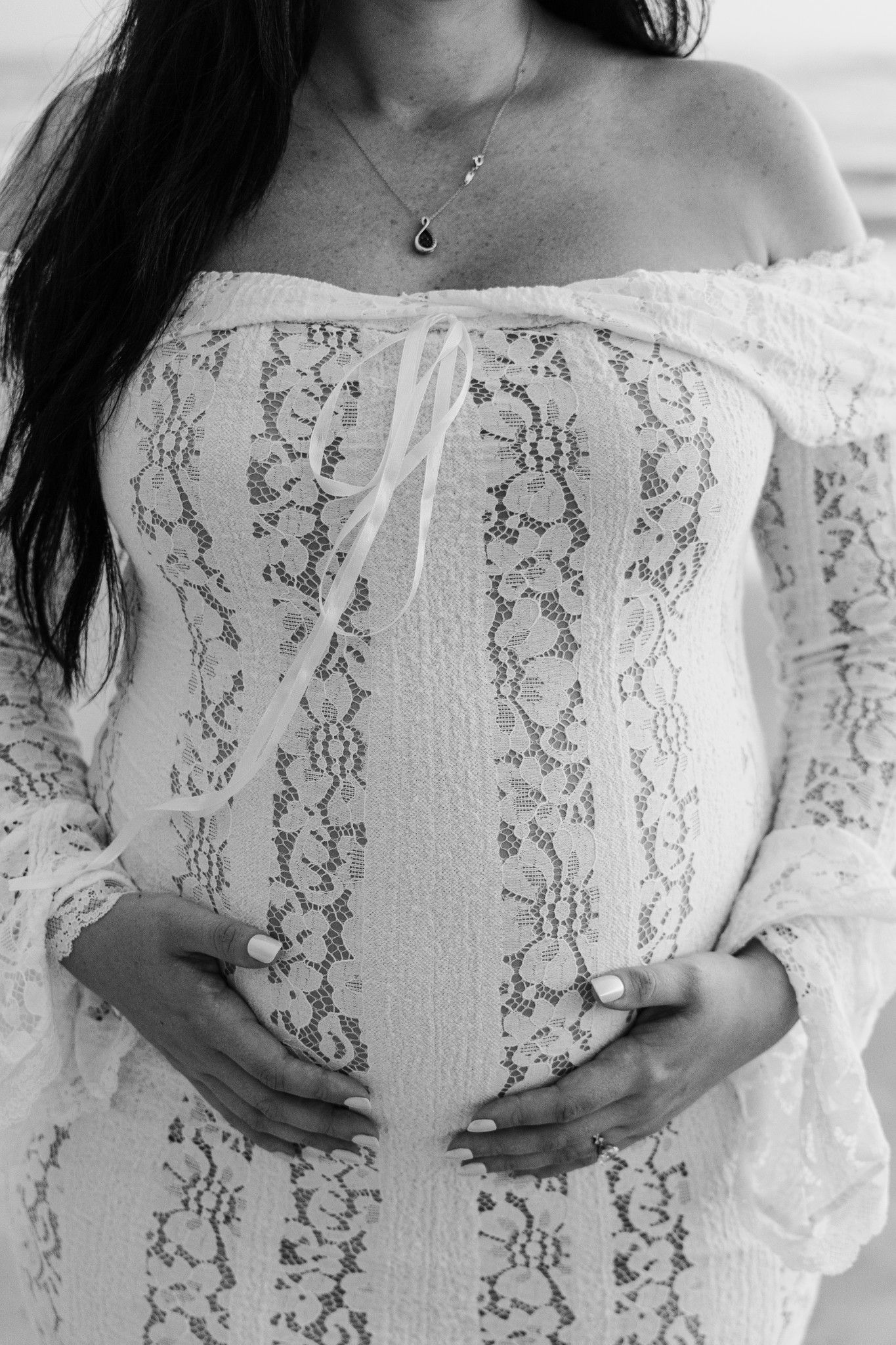 Black and white photo of pregnant woman in lace off-shoulder dress cradling baby bump with both hands.