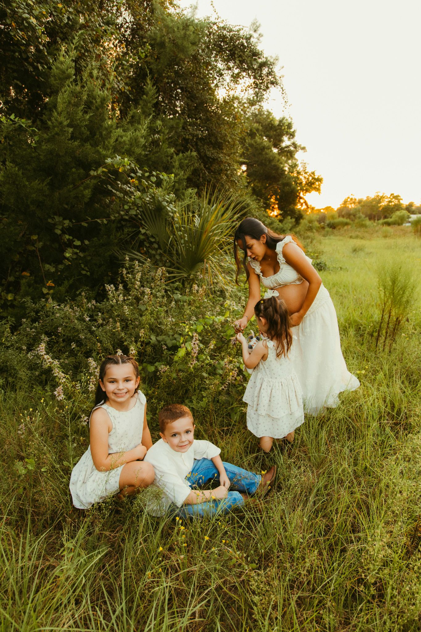 Family photo of mother and three children in white outfits playing in a lush green field at golden hour sunset.