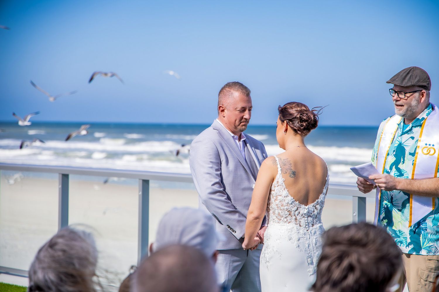 A small beach wedding ceremony takes place on the sand with guests gathered under a bright blue sky.