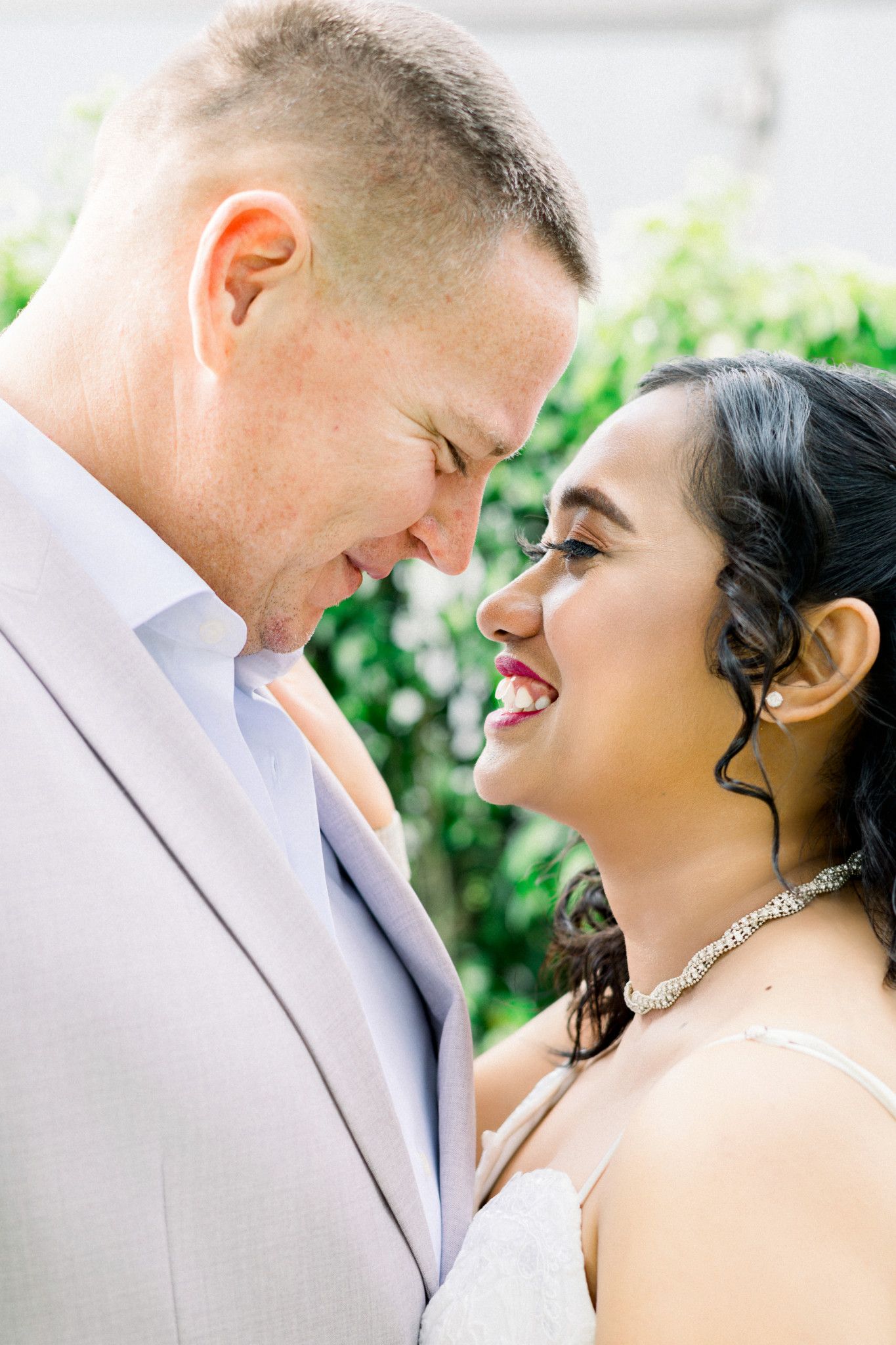 A smiling bride in a diamond necklace and groom in a gray suit share a romantic forehead-to-forehead moment outdoors.