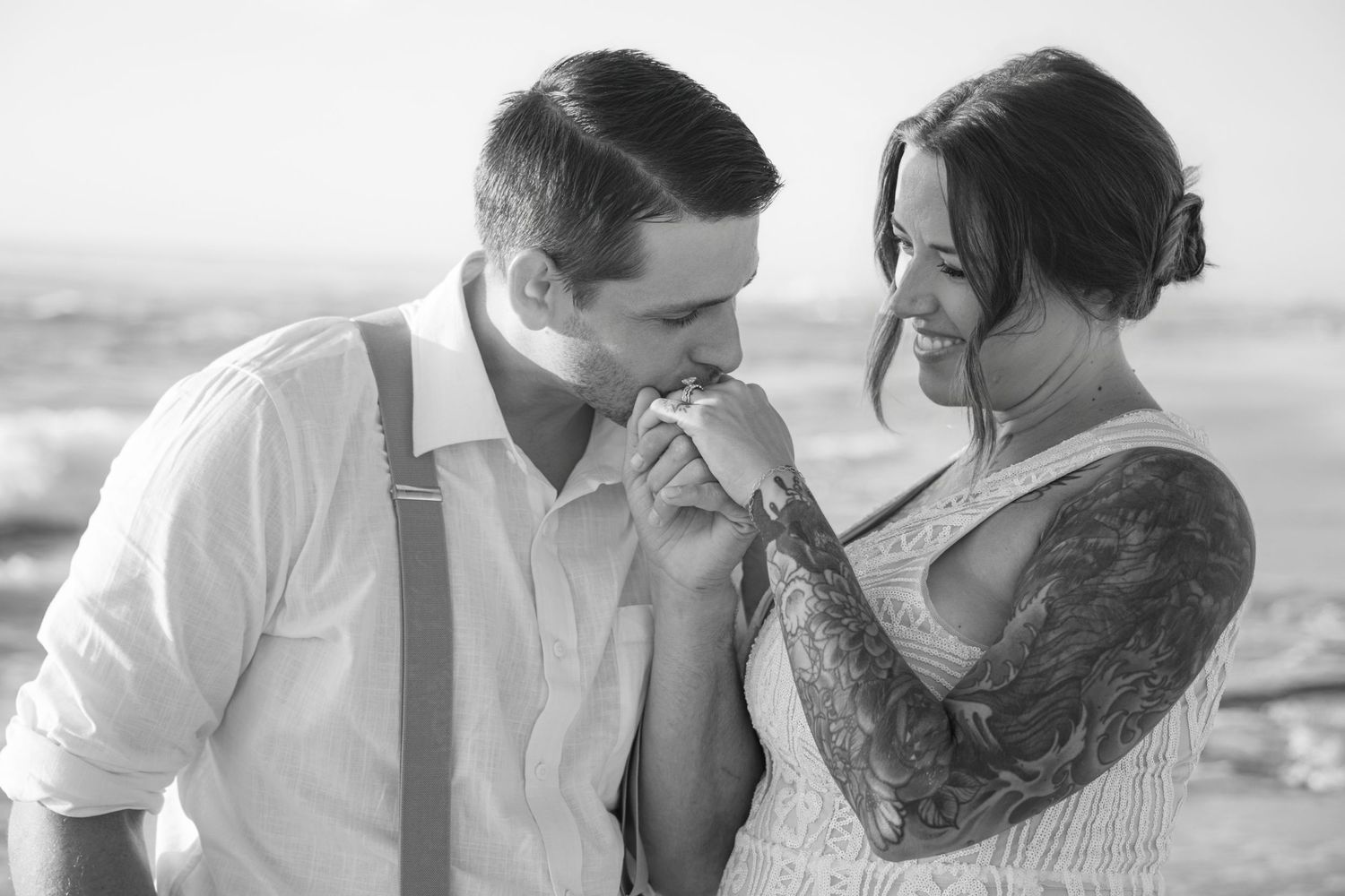 Black and white beach wedding photo of groom kissing bride's hand, bride smiling with tattoo sleeve visible.
