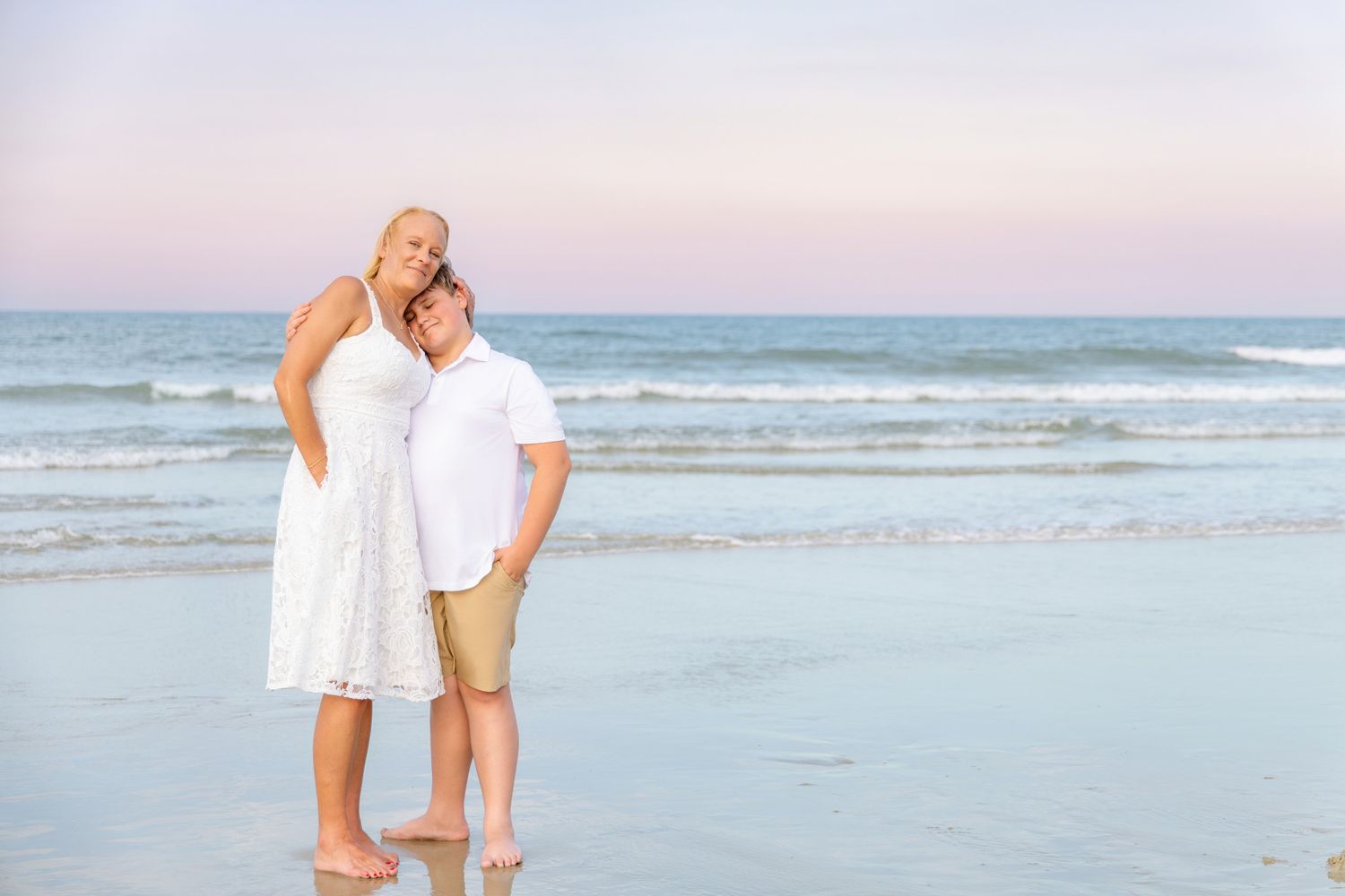 Couple embracing on a sandy beach at sunset, wearing white outfits, with calm ocean waves in the background.