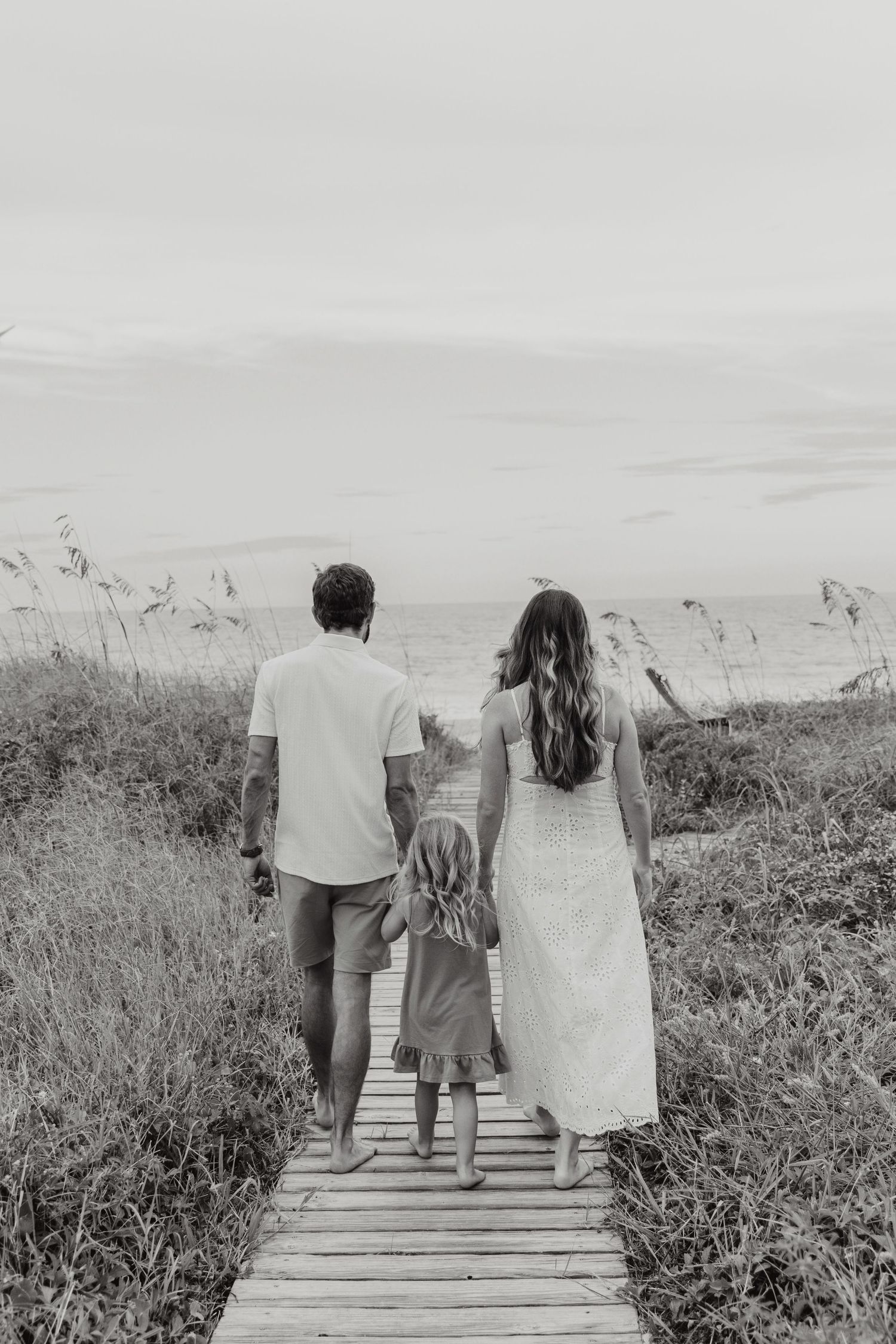 Black and white photo of a family walking hand-in-hand down a boardwalk path toward the ocean beach.