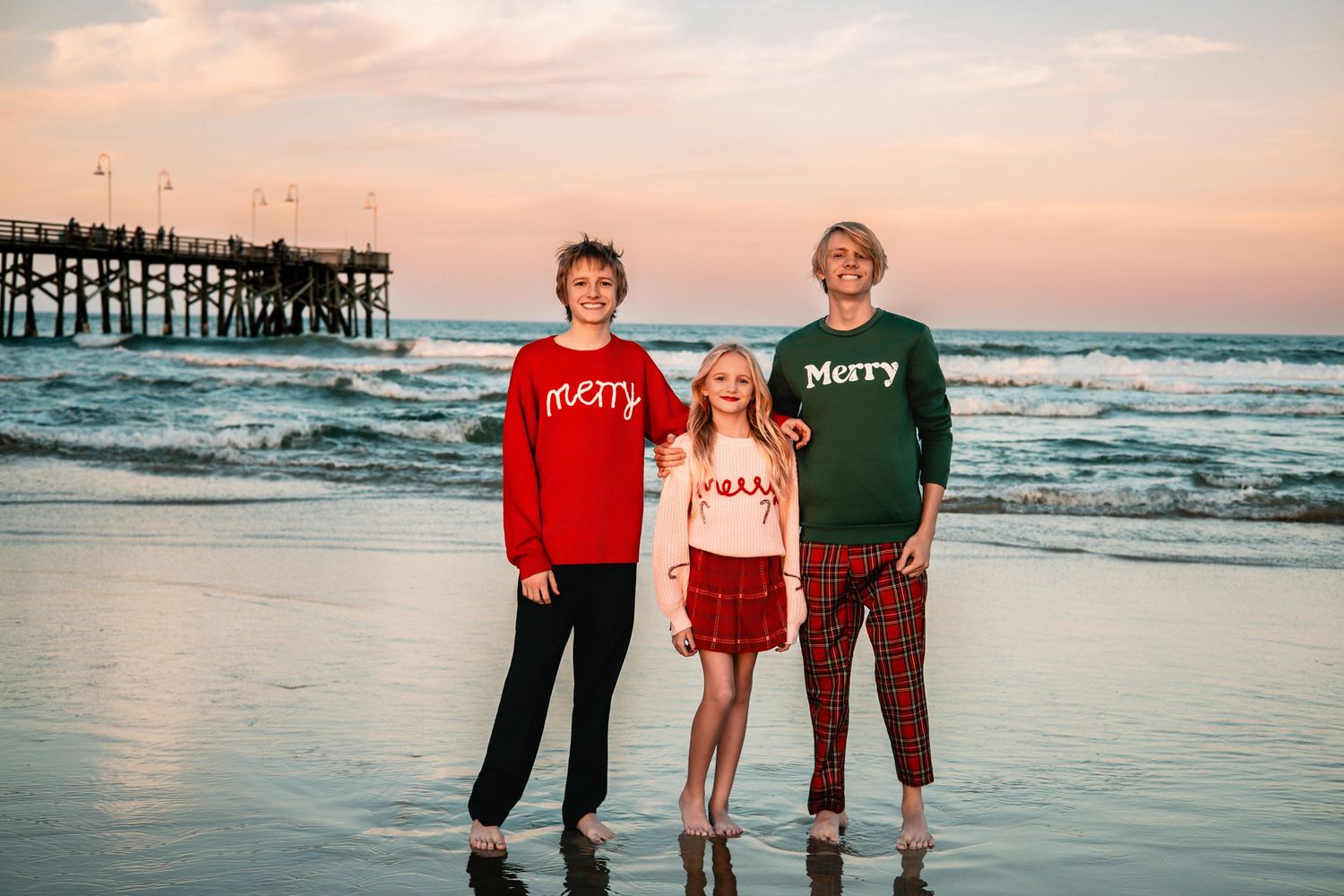 Group wearing festive red and green outfits poses on beach at sunset.
