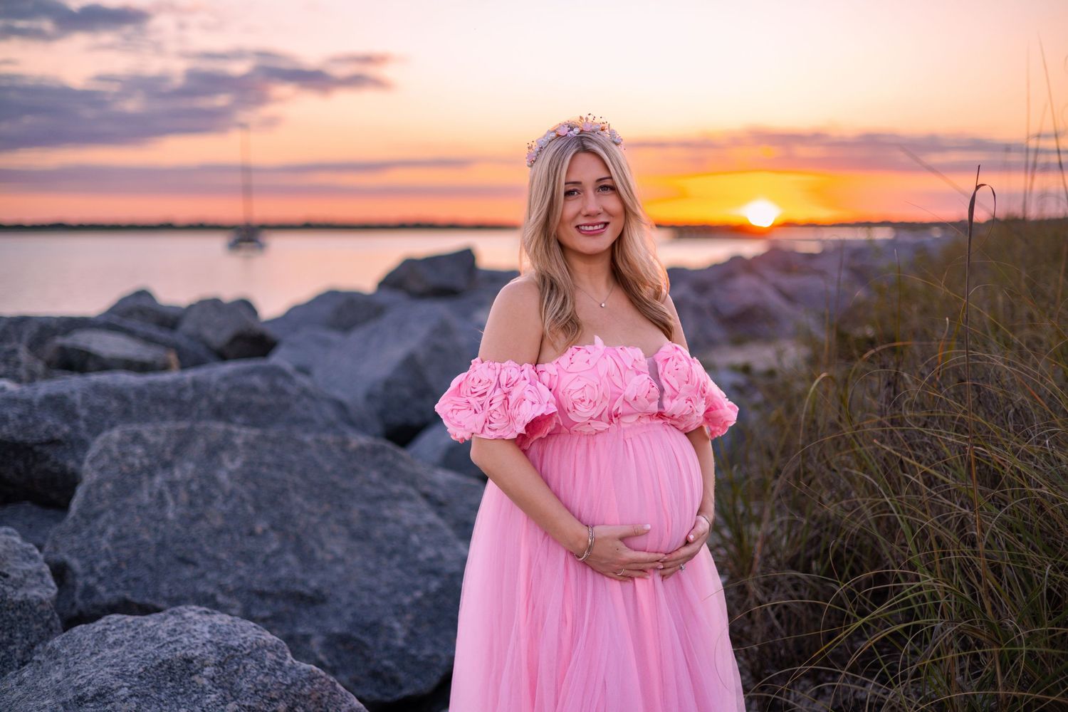 A woman in a flowing pink dress poses on a rocky beach during golden hour sunset.