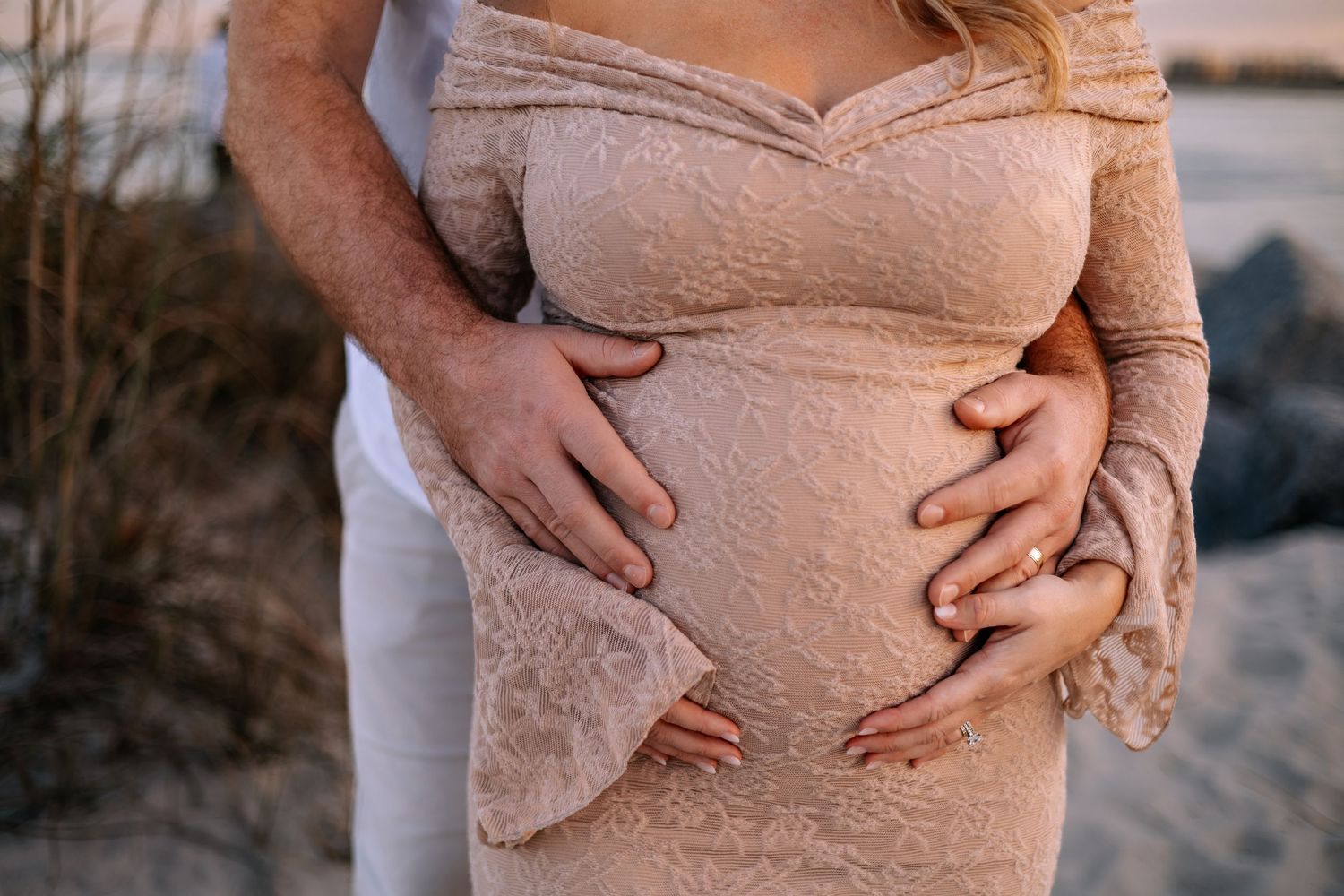 Close-up of a pregnant belly in a lace dress being held by loving hands on a beach at sunset.