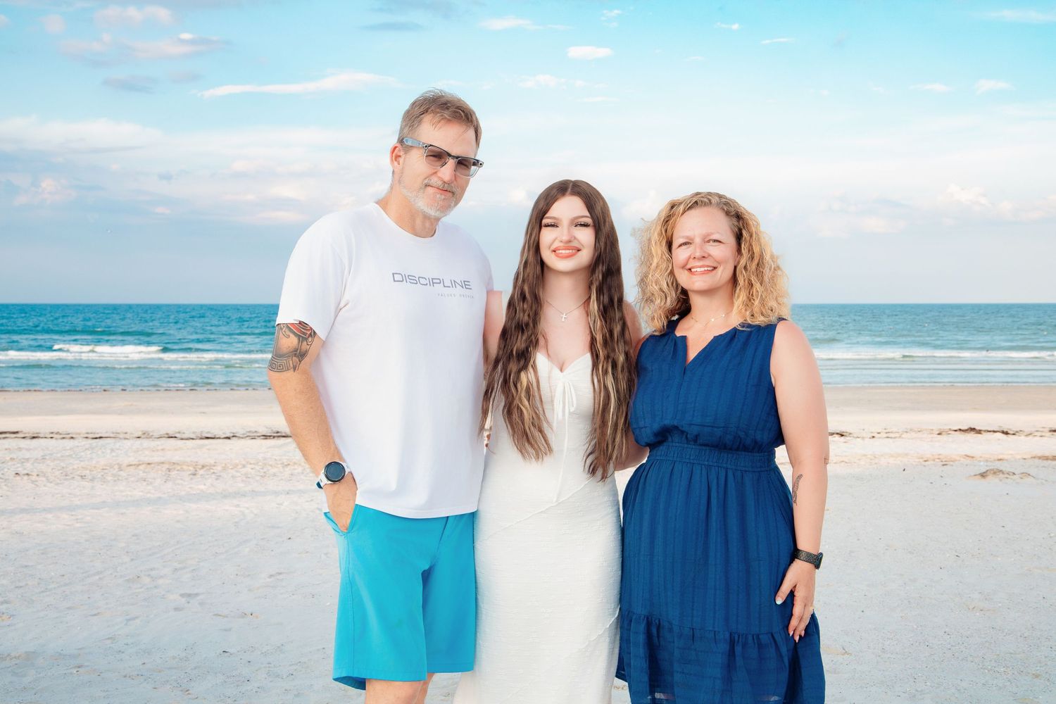 Family of three smiling together on a sunny beach, woman in white dress flanked by man in turquoise shorts and woman in blue dress.
