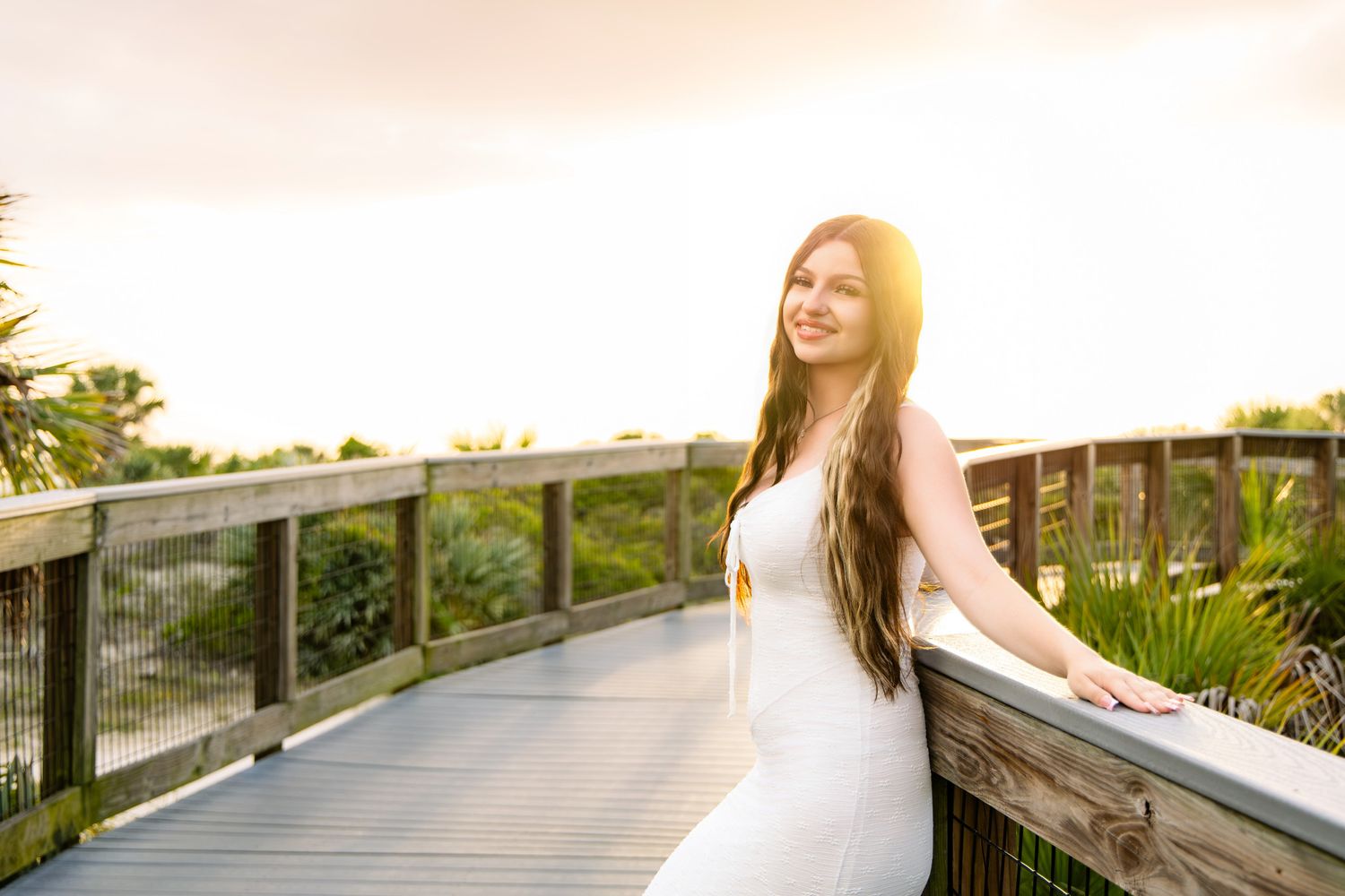 Smiling woman in white dress on wooden boardwalk surrounded by lush greenery at sunset.