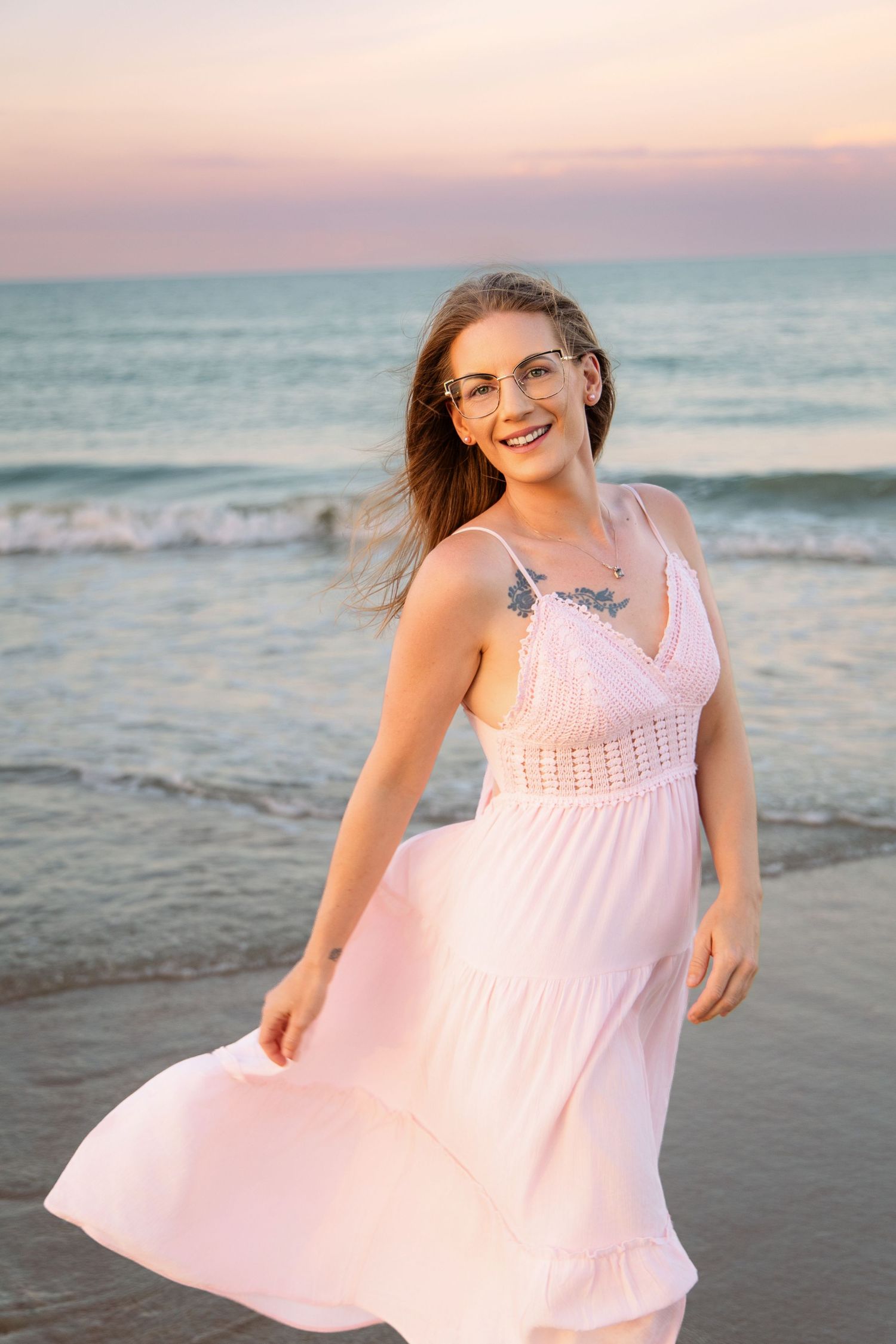 Young woman in pink sundress and glasses smiling on beach at sunset with ocean waves behind her.