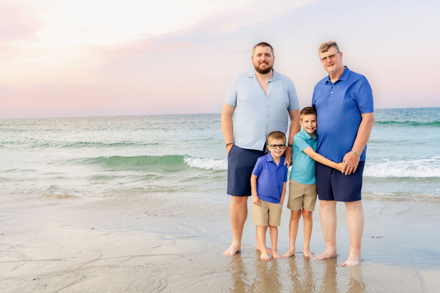 Two men and two boys pose together on a sandy beach at sunset with ocean waves behind them.