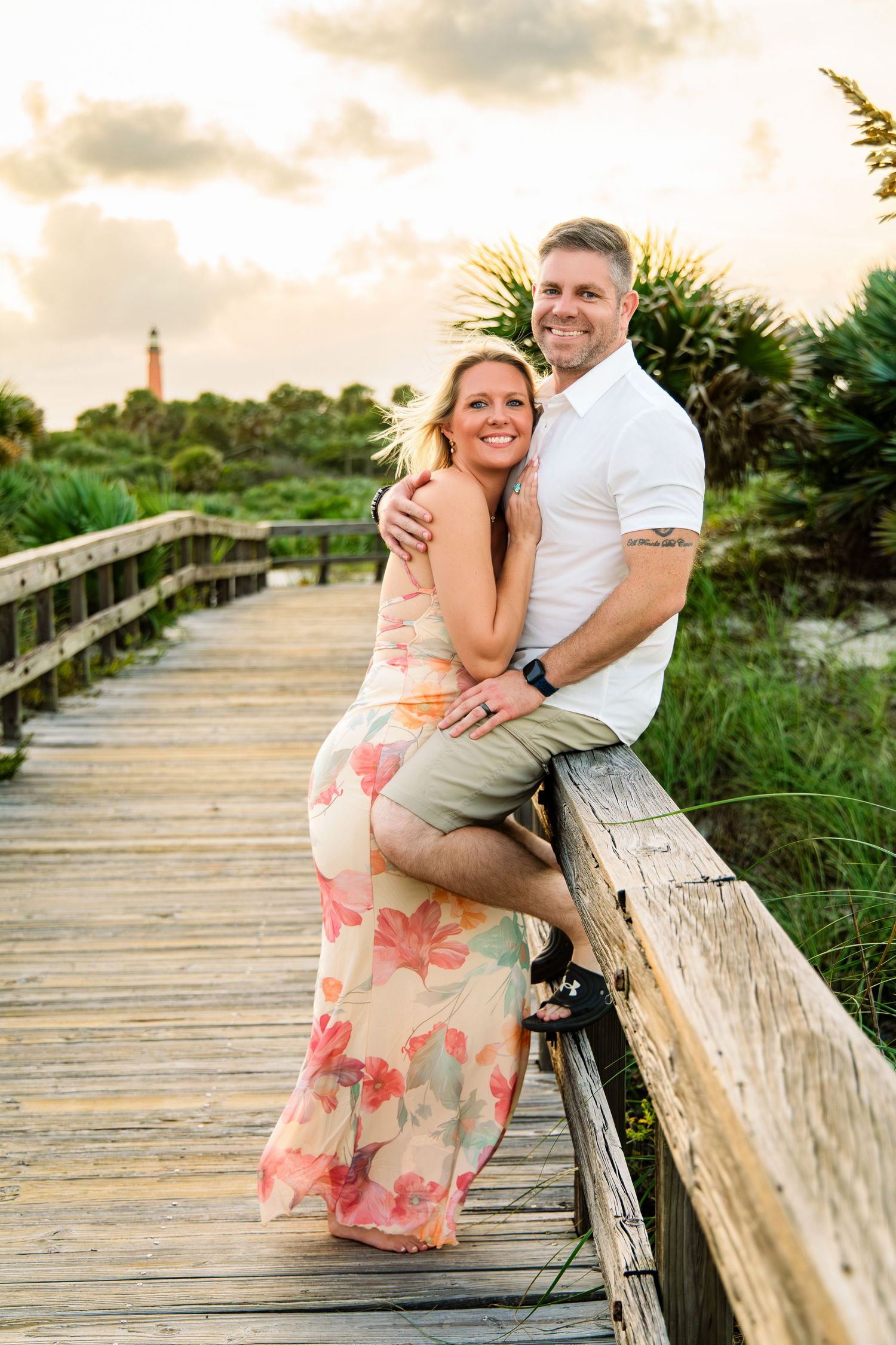 Couple embracing on a wooden boardwalk near a lighthouse at sunset, woman in floral dress, man in white shirt.