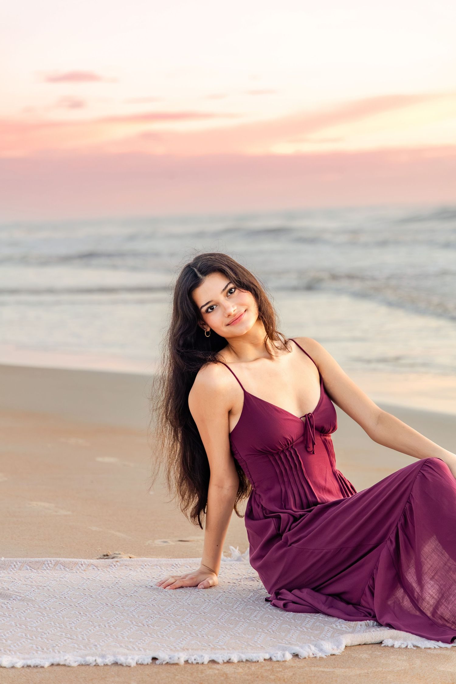Young woman in burgundy maxi dress sitting on beach blanket at sunset, smiling at camera with ocean waves behind her.