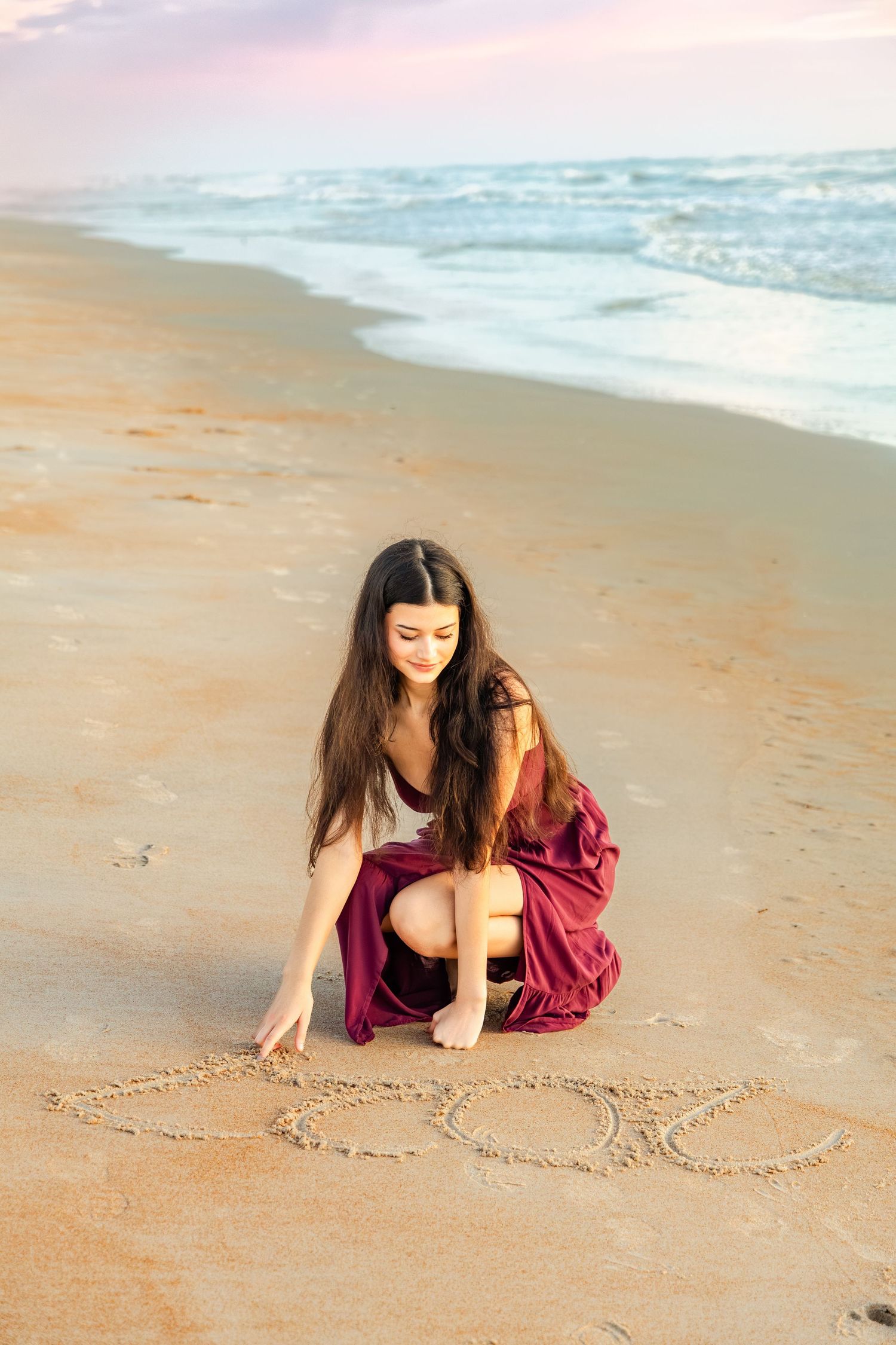 Young woman in burgundy skirt writing in wet sand on a beach at sunset with ocean waves in background.