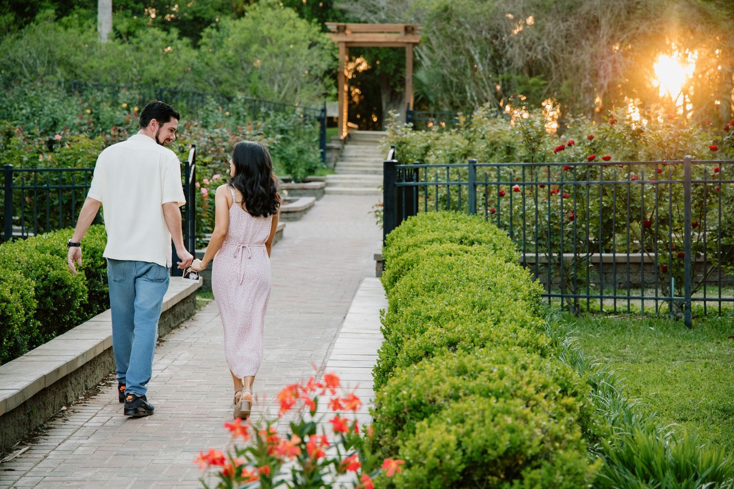 Couple walking hand in hand along a garden pathway surrounded by lush greenery and blooming flowers at sunset.