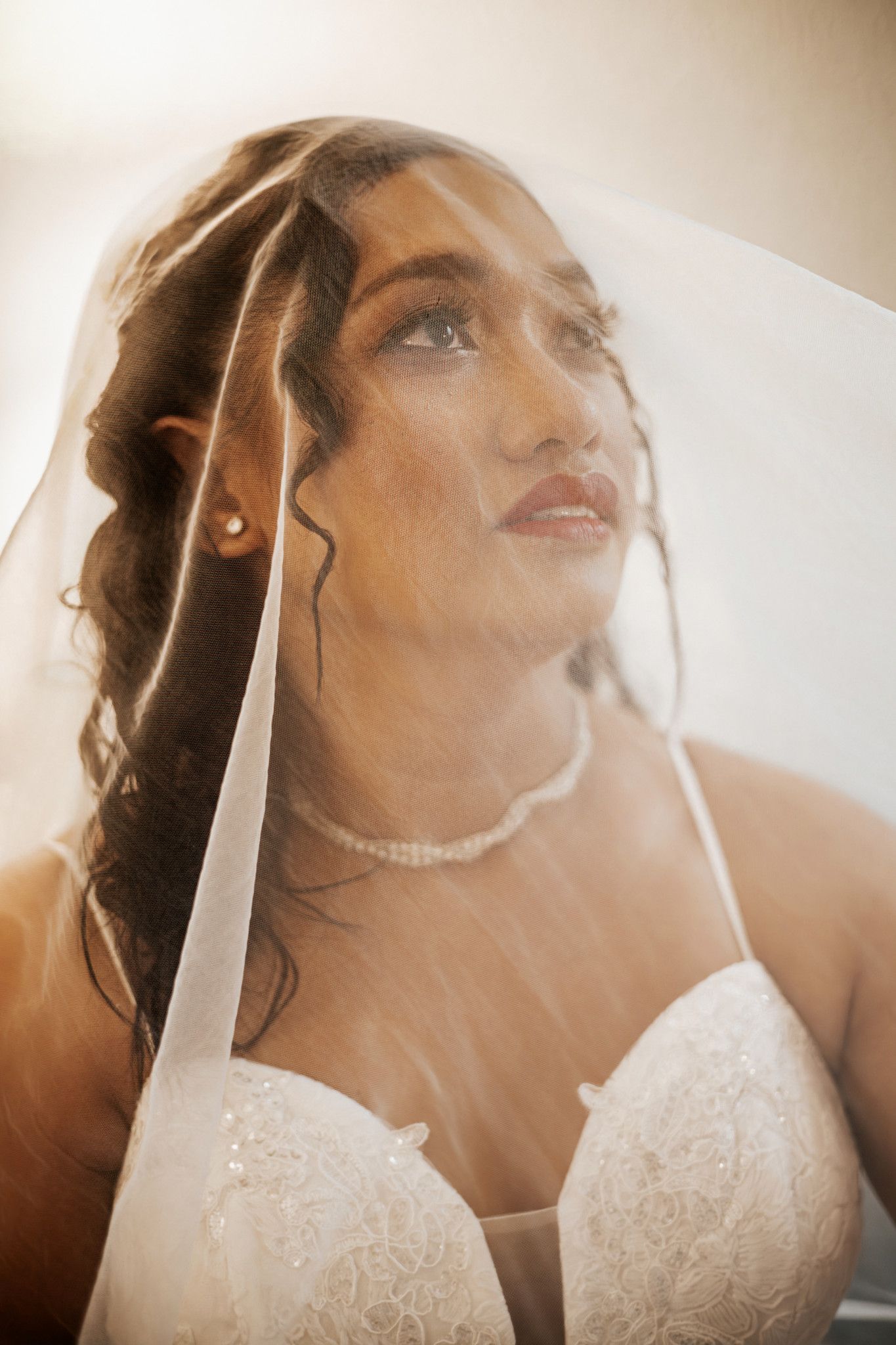 Bride in lace wedding dress and veil with pearl necklace gazing upward in soft natural light.