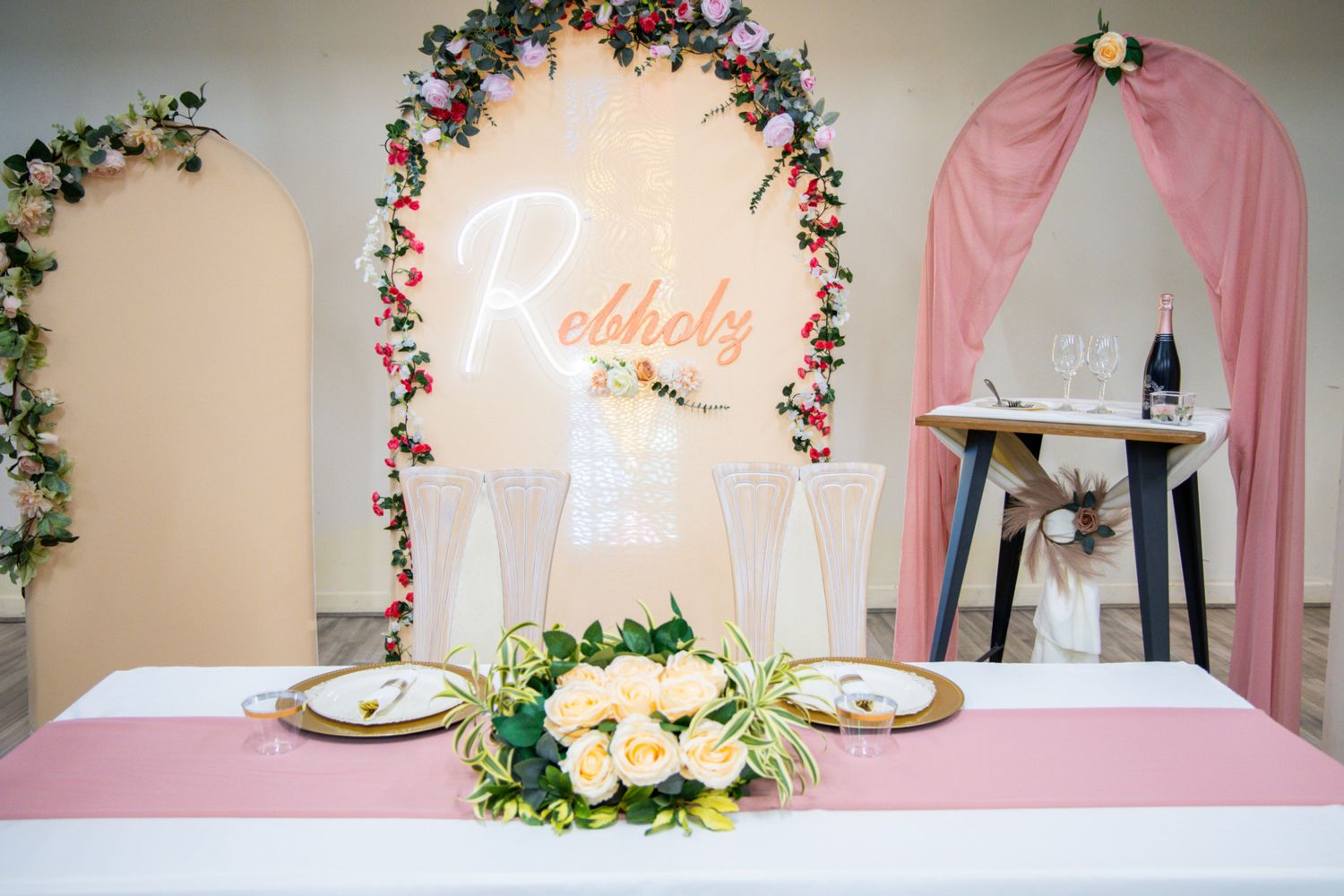 Elegant wedding sweetheart table with floral arch, pink draping, gold chargers, and glowing neon name sign.