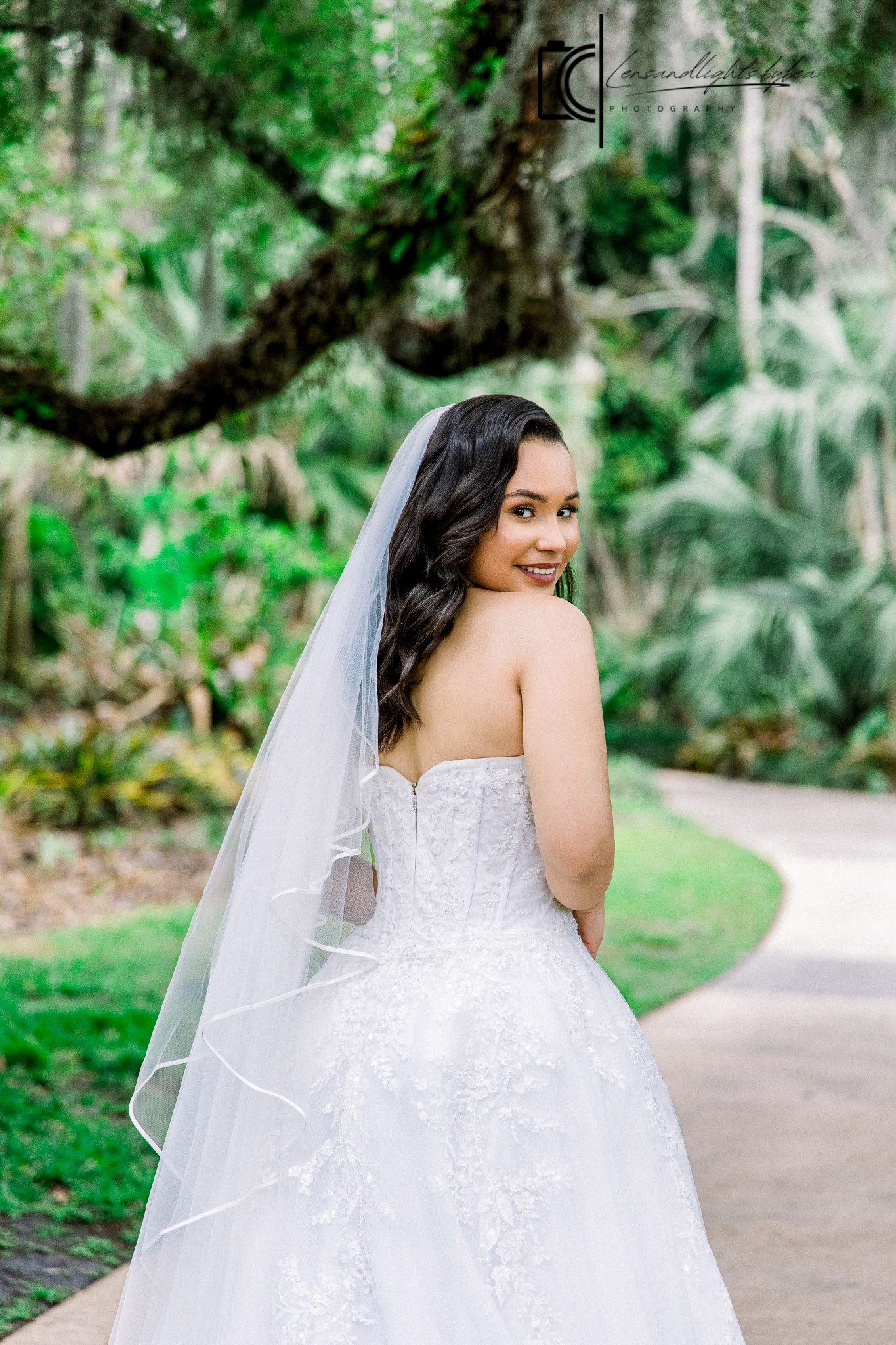 Bride in strapless lace ballgown with veil poses gracefully on garden path surrounded by lush greenery.
