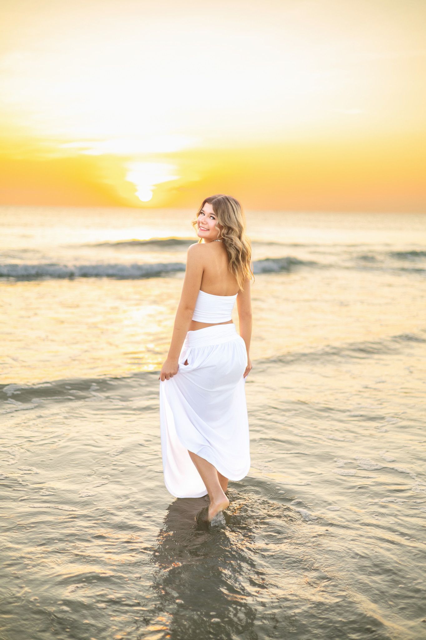 Young woman in white maxi skirt walks in ocean waves at golden sunset beach.