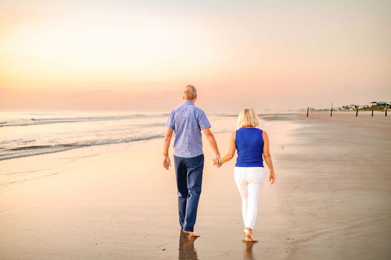 An elderly couple holds hands while walking barefoot on a beach at sunset, enjoying a romantic stroll by the ocean.