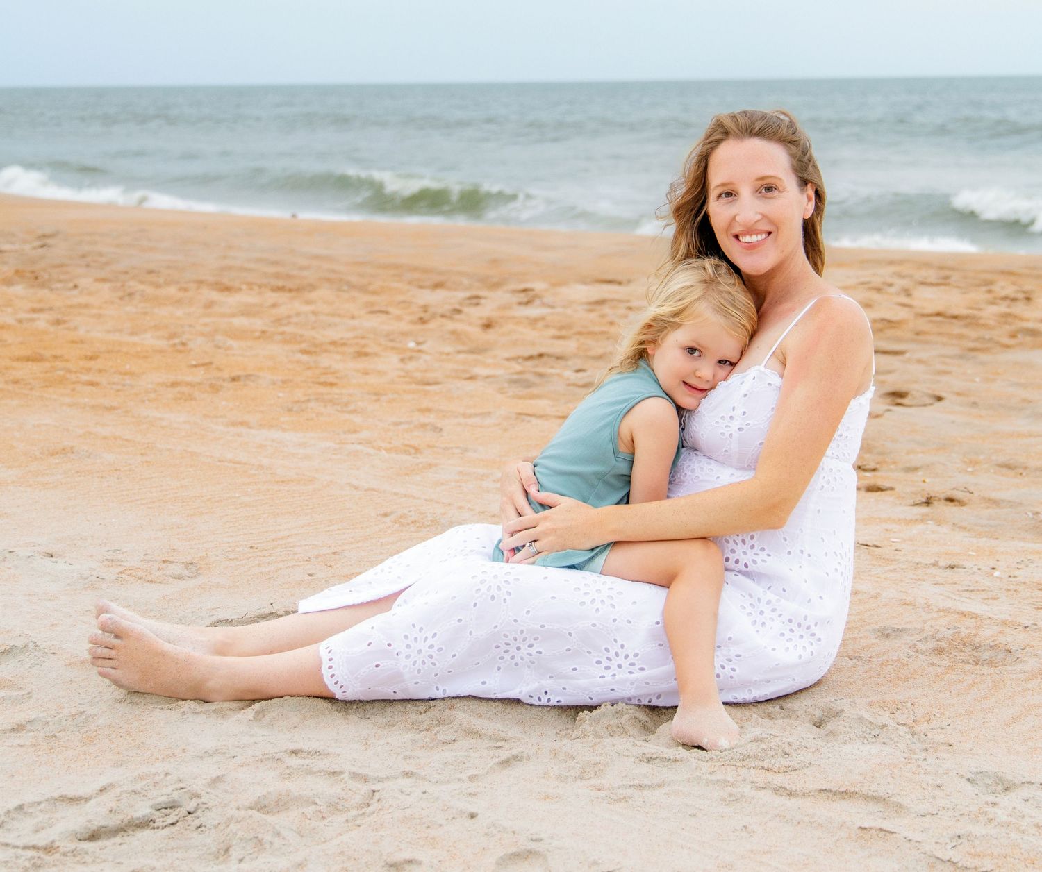A smiling woman in a white dress sits on a sandy beach hugging a young child, with ocean waves in the background.