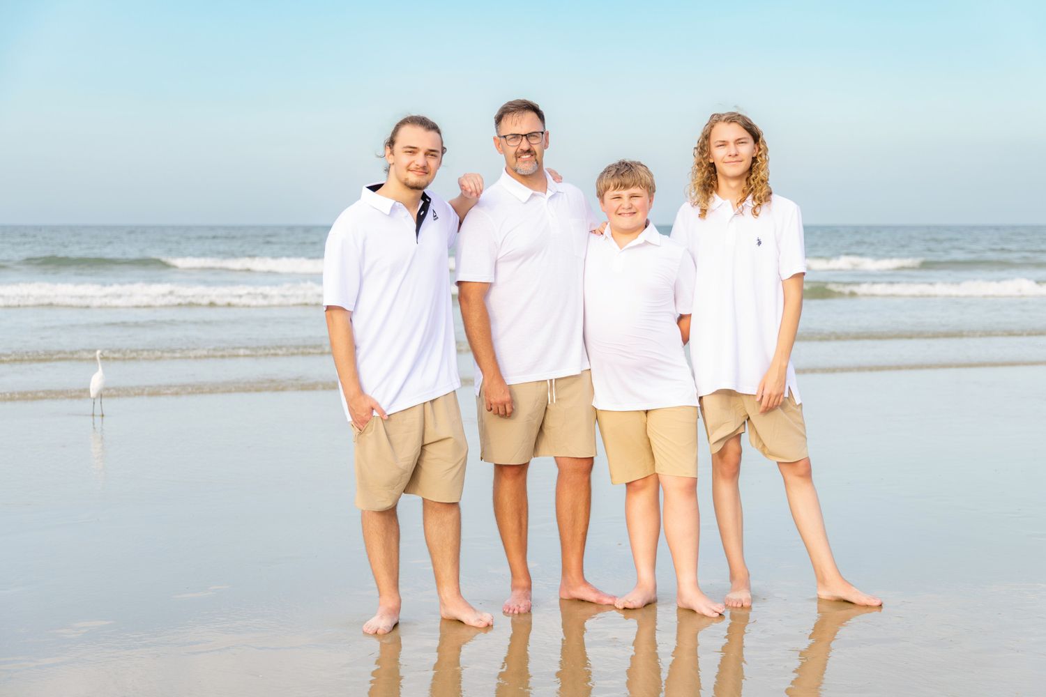 Family of four in matching white shirts and khaki shorts posing together on a sunny beach with ocean waves behind them.