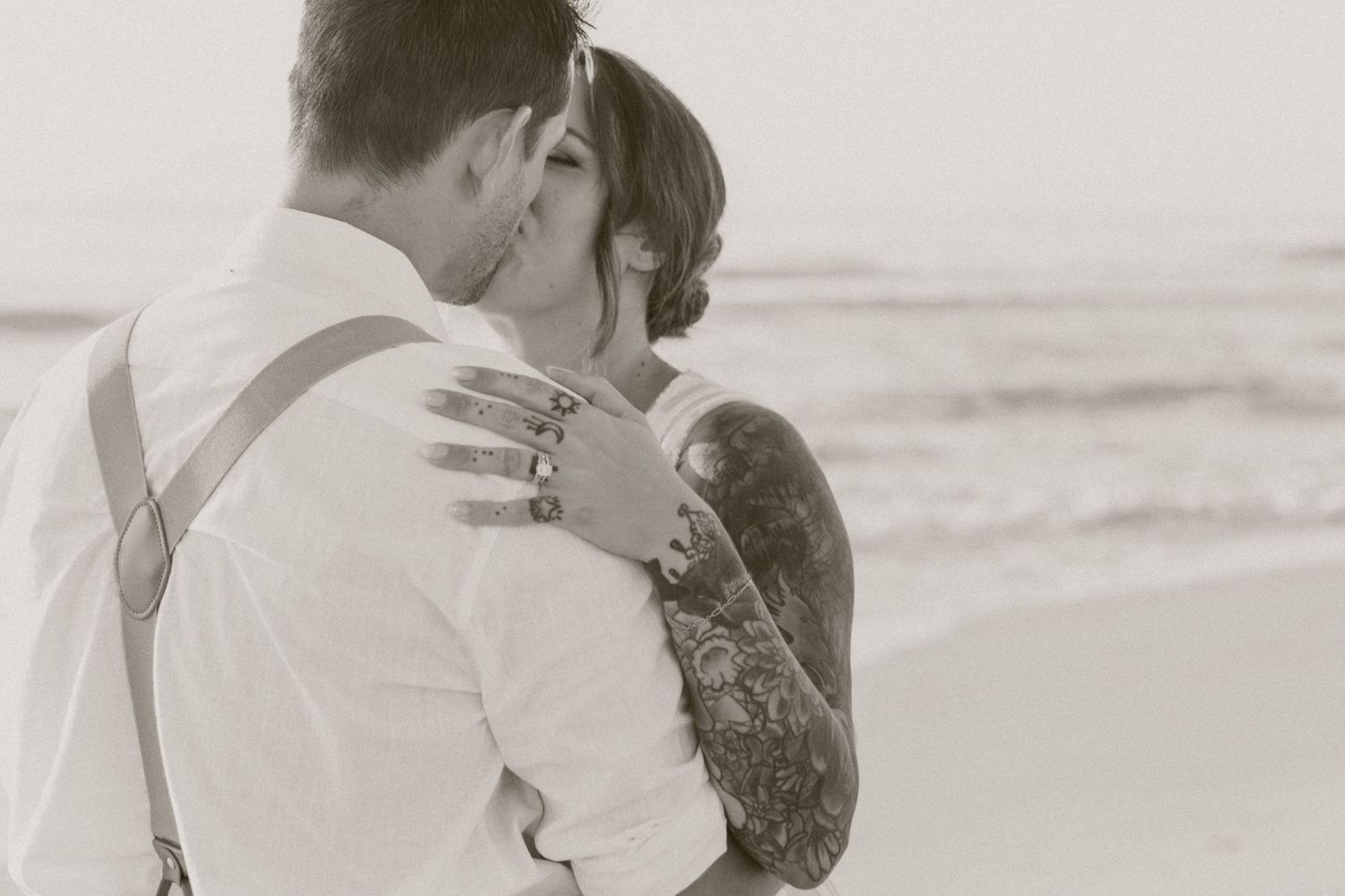 Black and white photo of couple kissing on beach, bride's tattooed arm around groom.