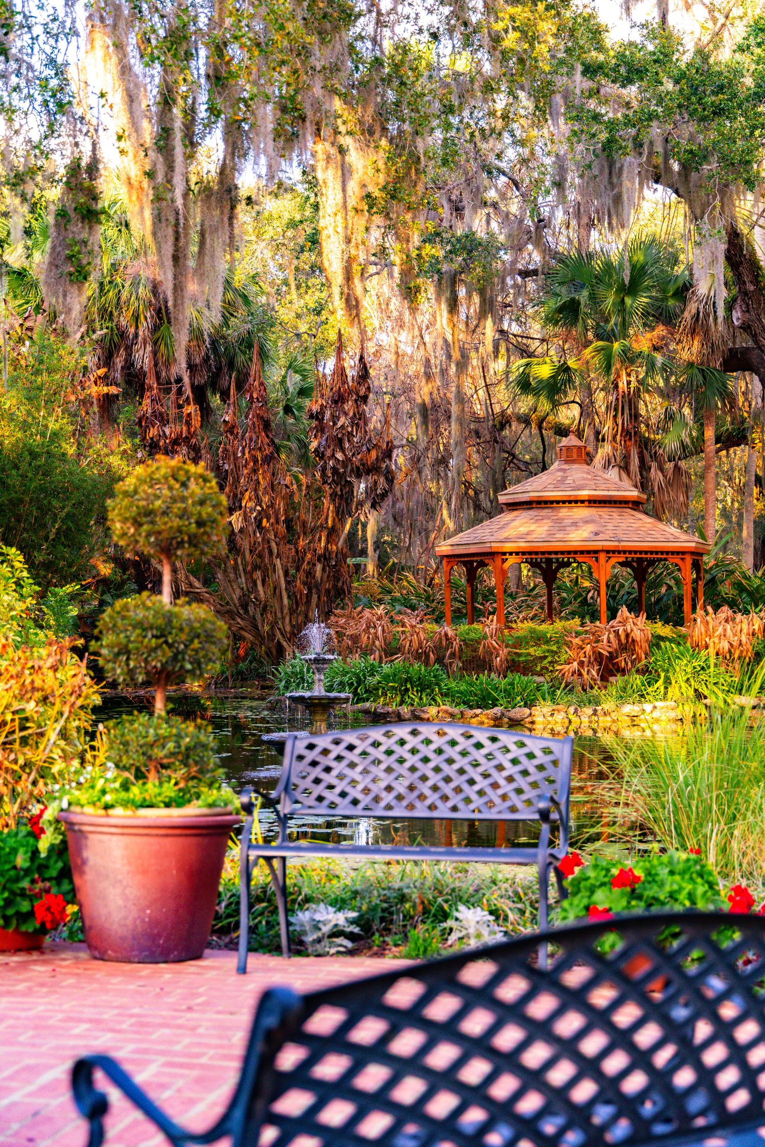 Lush garden patio with iron bench, gazebo, and pond surrounded by Spanish moss-draped trees and colorful flowers.