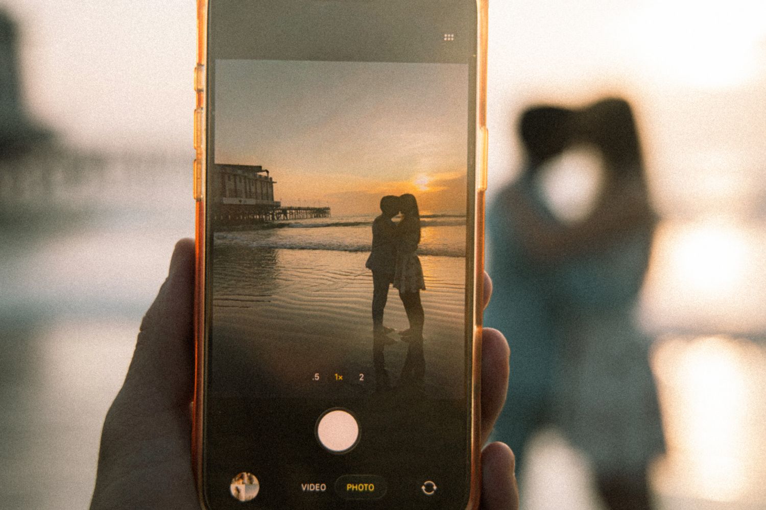 A hand holds a smartphone capturing a couple kissing on a beach at sunset near a pier.