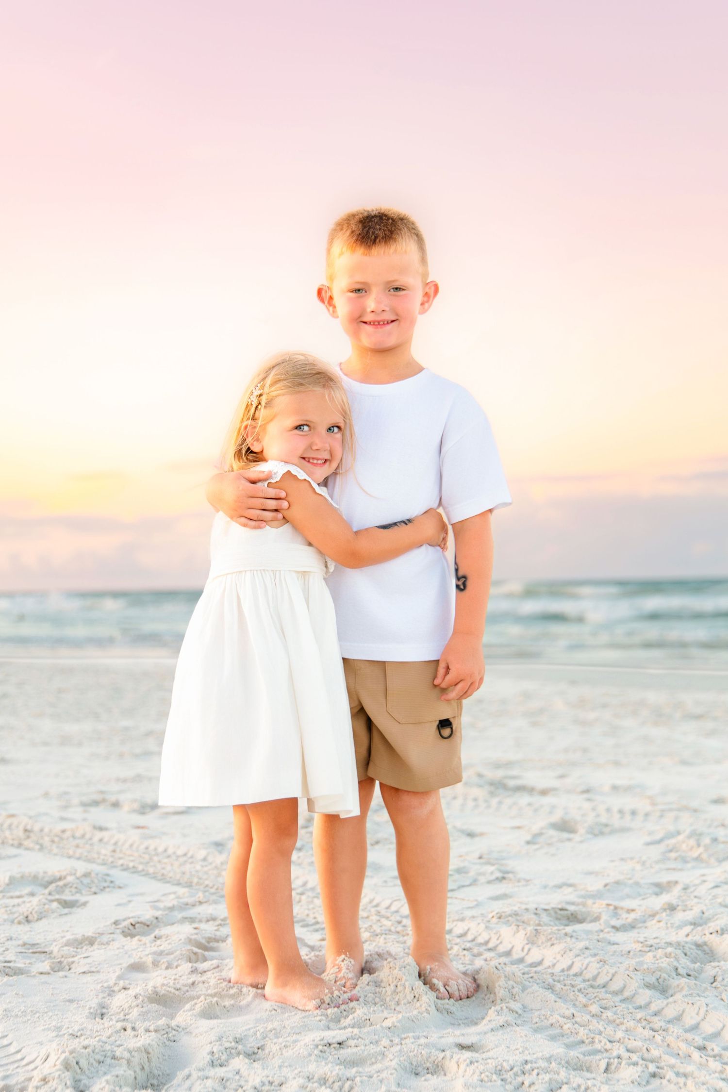 Two young siblings hug and smile on a sandy beach at sunset, girl in white dress and boy in white shirt.