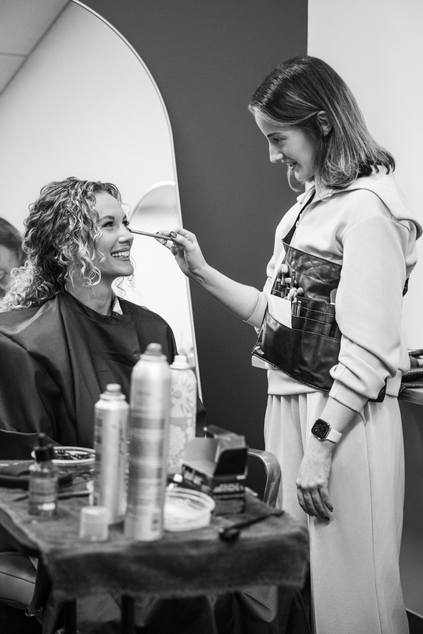 Black and white photo of a makeup artist applying cosmetics to a smiling woman in a salon chair.