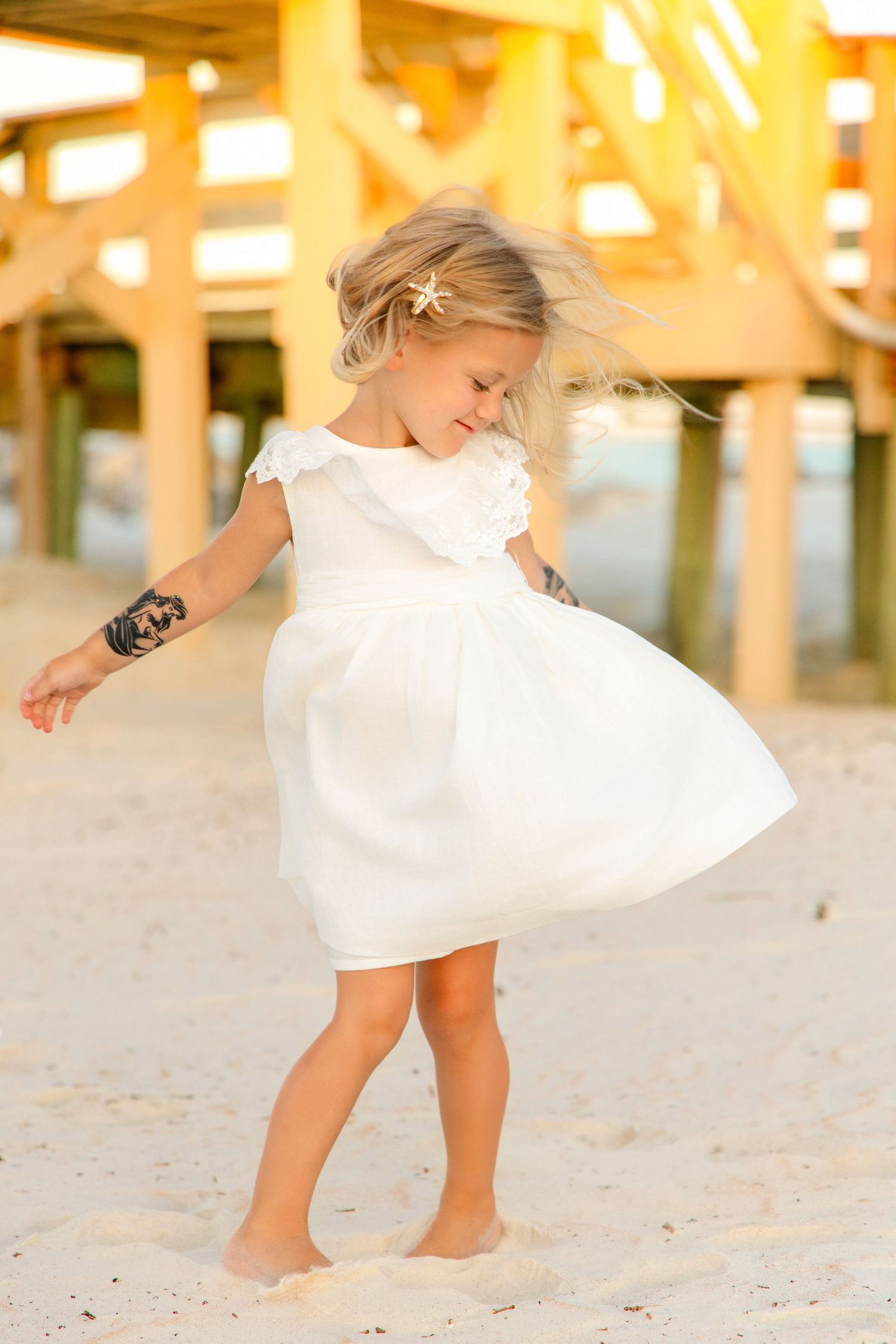 A young girl in a white dress twirls barefoot on sandy beach near pier at sunset.