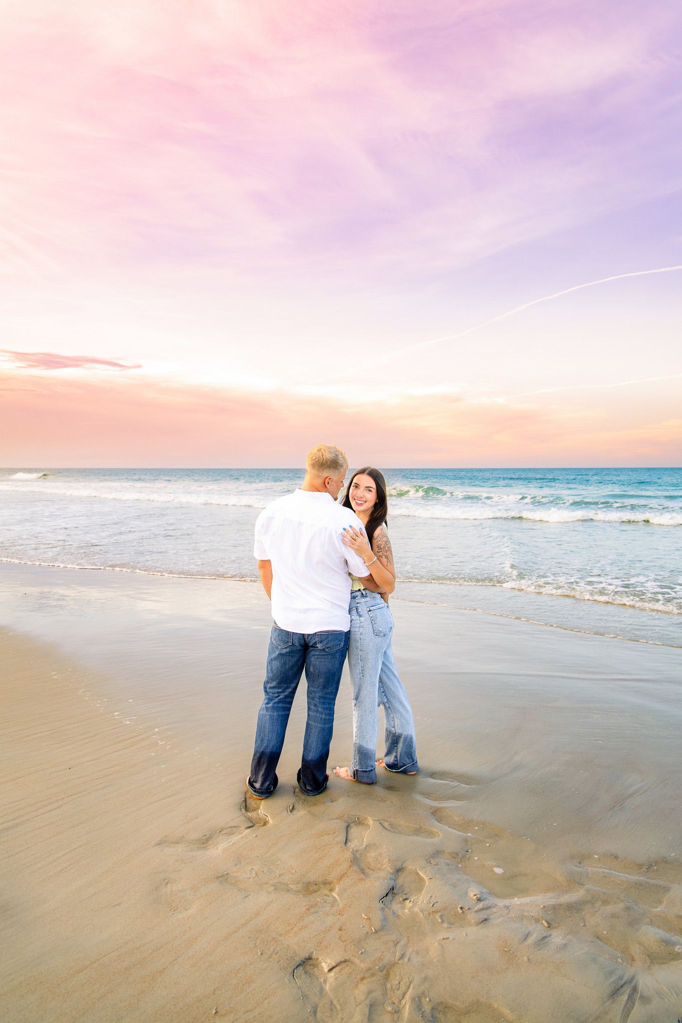 Couple embracing on a sandy beach at sunset with colorful pink and purple sky and ocean waves in the background.