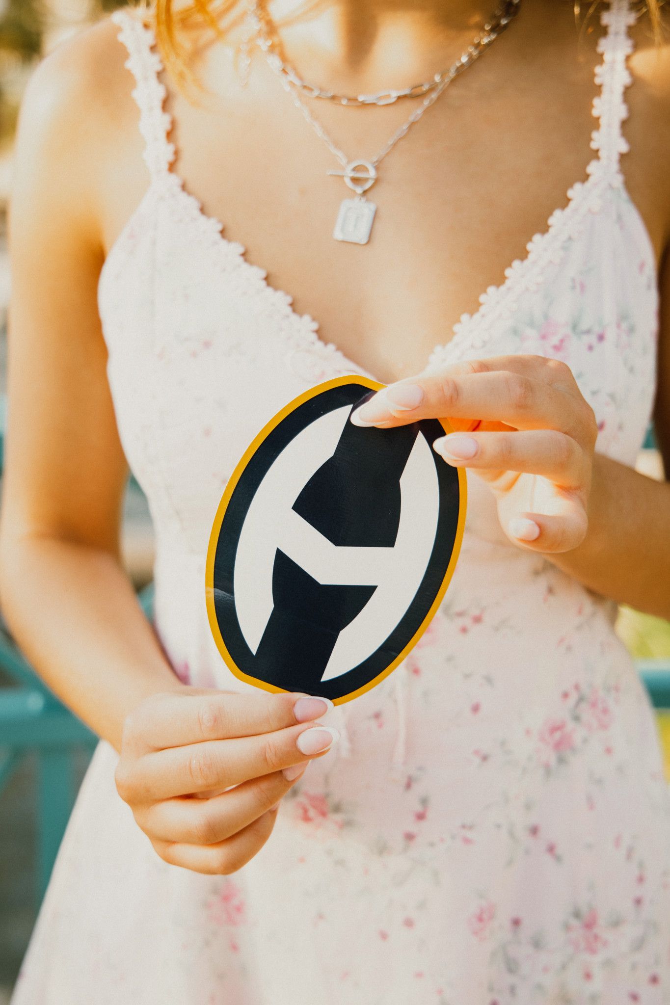 Woman in floral dress holding a black and yellow oval logo sticker with white manicured nails.