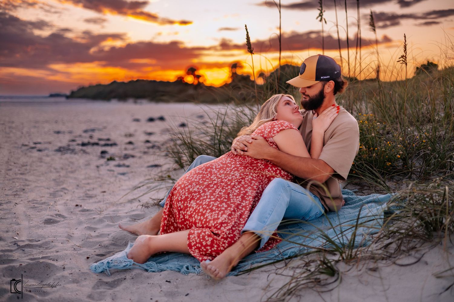 Romantic couple lying on beach blanket during vibrant orange sunset with coastal grasses in foreground.