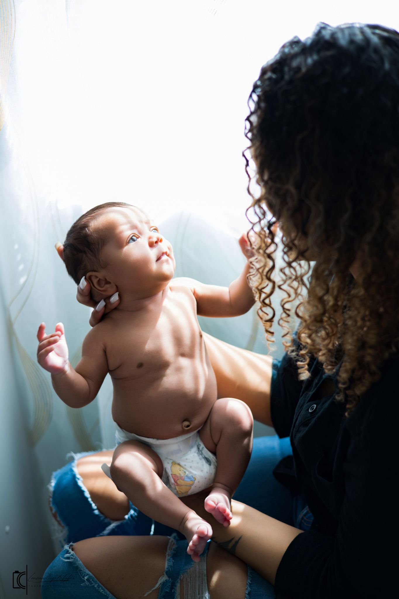 Intimate portrait of a baby in a diaper reaching up while being held against a bright window.