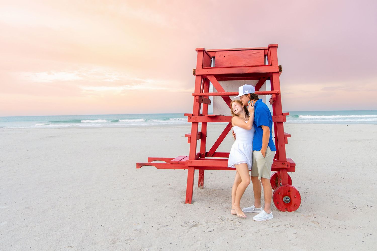 Couple embracing by a red lifeguard stand on a sandy beach at sunset with a colorful pink sky.