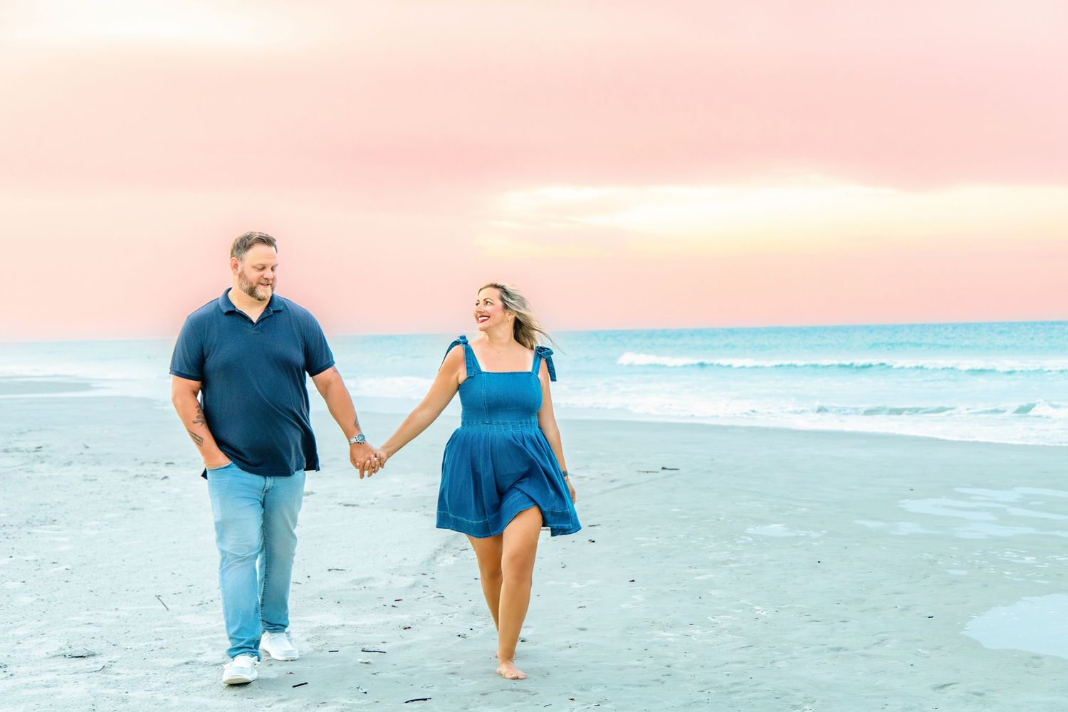 Couple holding hands walking on beach at sunset with pink sky and ocean waves in background.