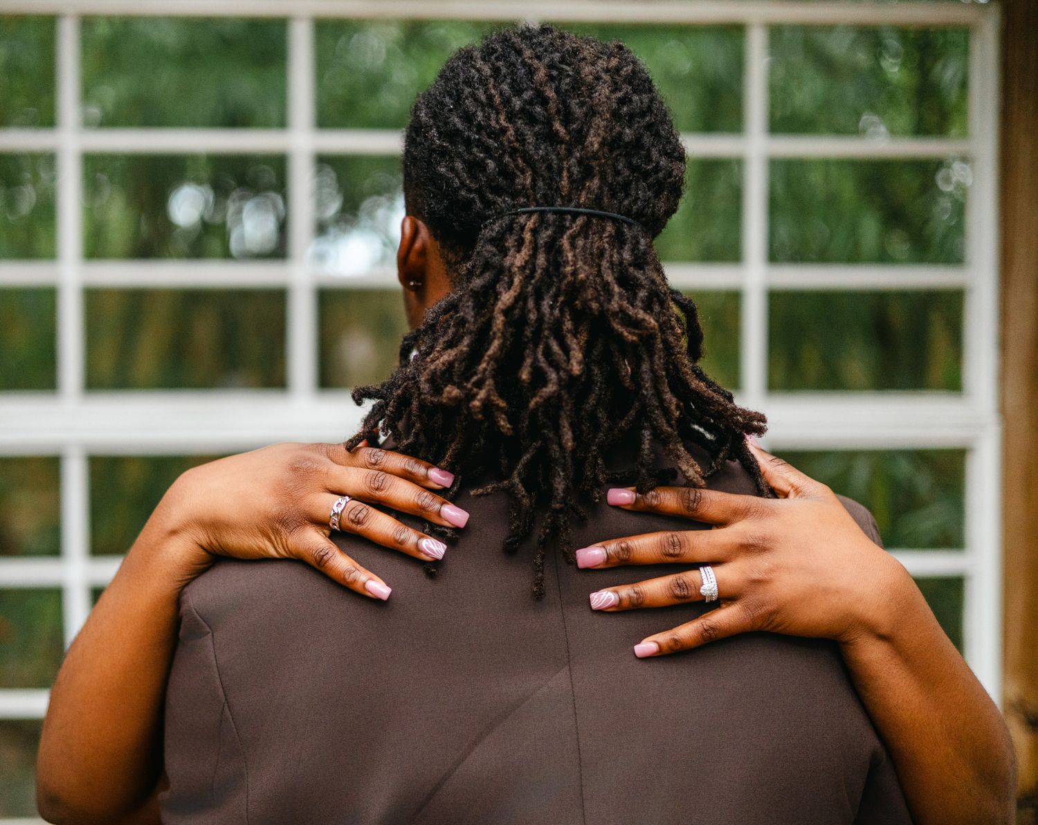 A person with braided hair wearing a brown shirt is shown from behind against a white fence background.